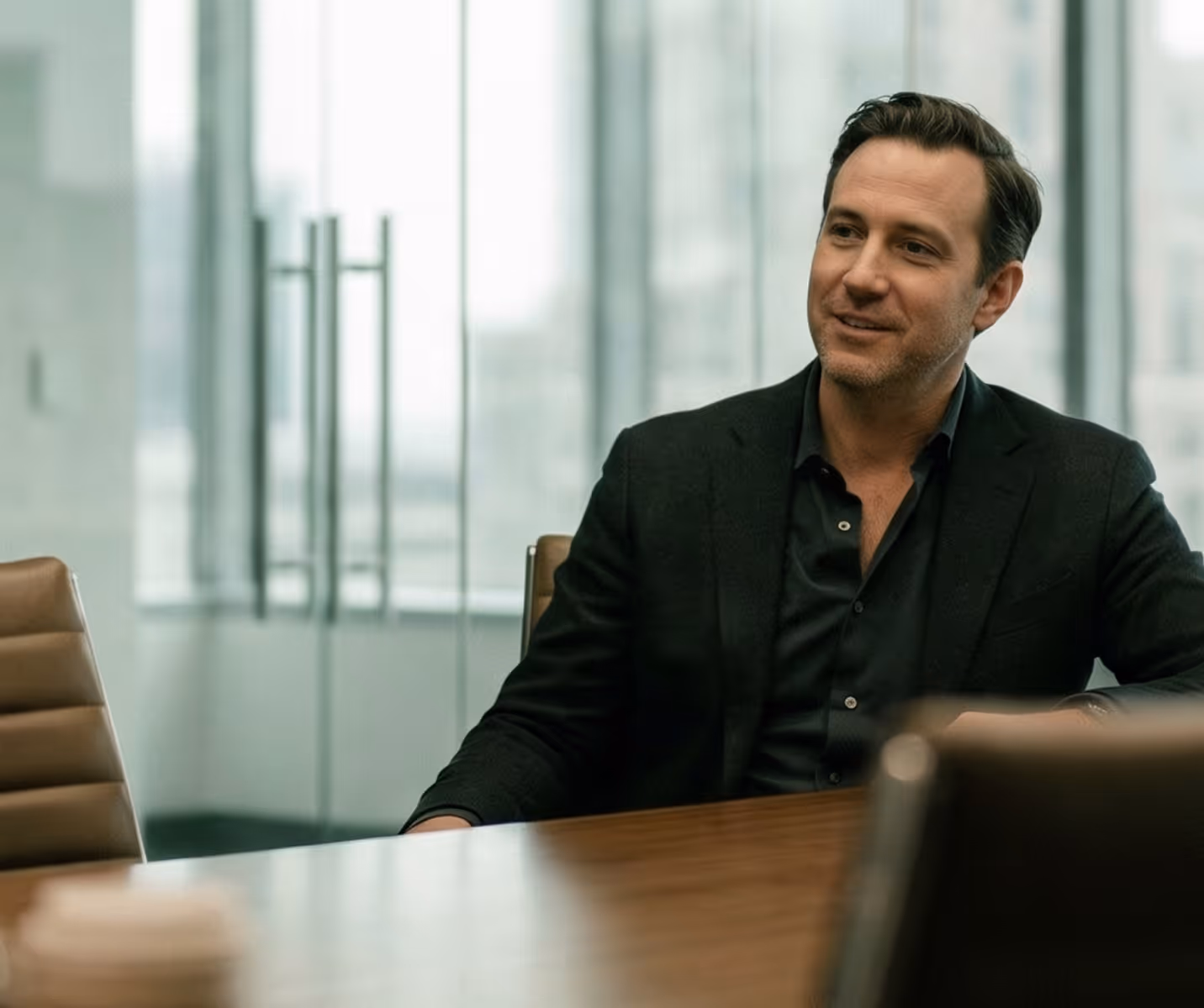 Man in a black suit sitting at a conference table in a modern office with glass walls.