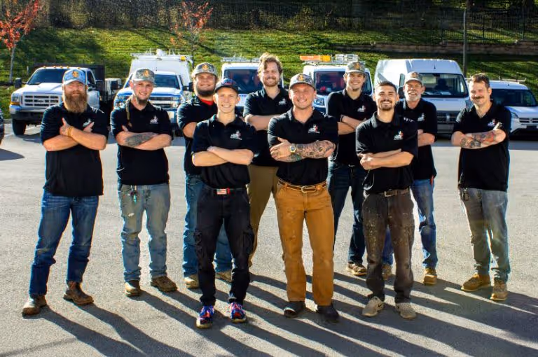 Group of ten men wearing black shirts and caps standing with arms crossed in a parking lot with white work vans behind them.