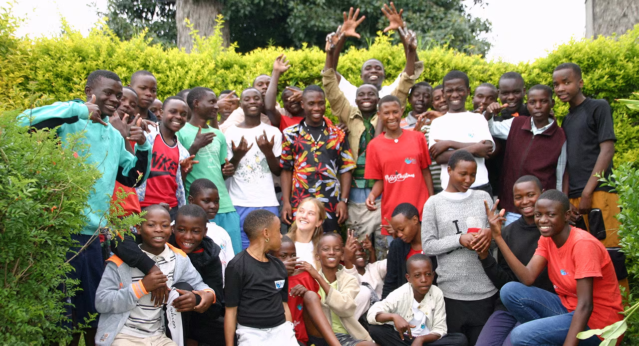 Group of smiling children and teenagers posing outdoors with greenery in the background.