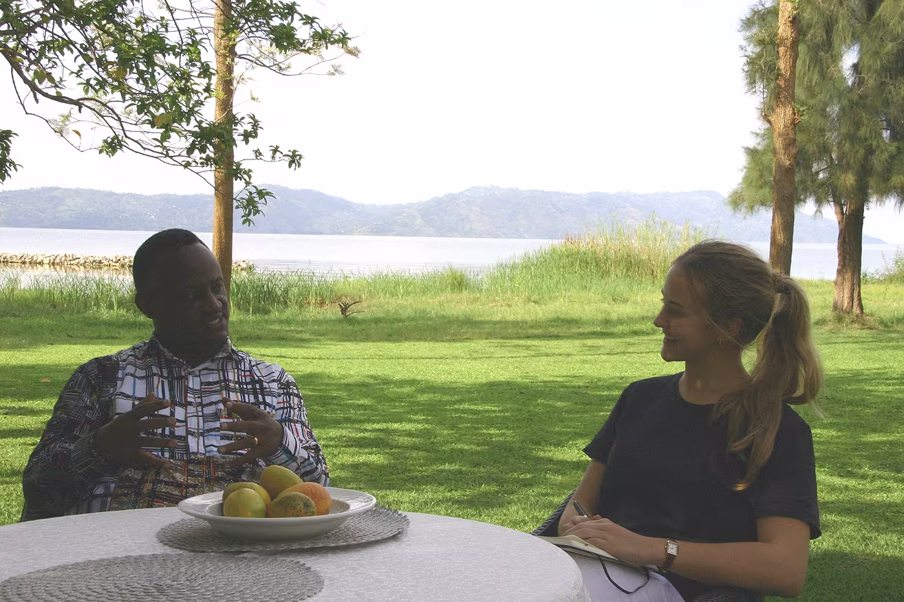 Two people sit at a round outdoor table with fruit, talking on a grassy lawn beside a lake with trees and hills in the background.