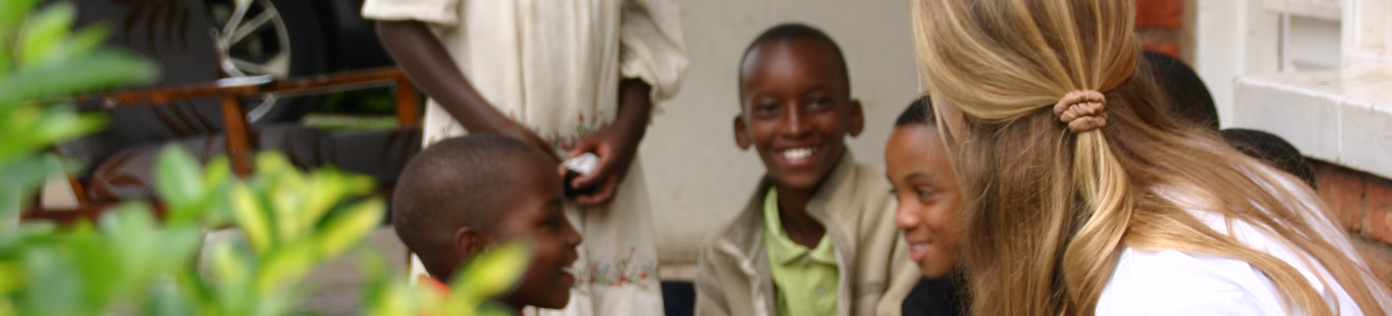 A woman crouches and smiles while talking with a small group of children sitting outside near a brick wall and greenery.