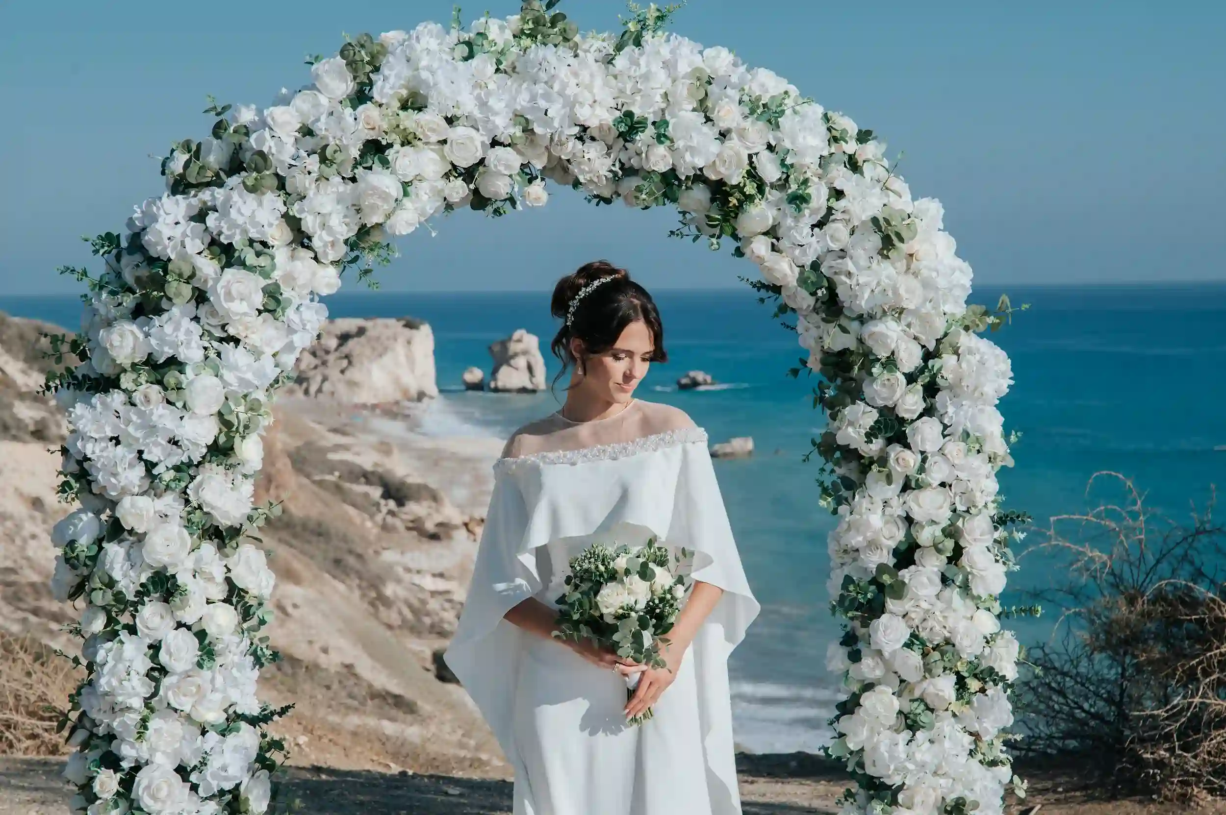 כלה עומדת תחת קשת פרחים מול הים במהלך חתונת יעד רומנטית בקפריסין לזוג מישראל