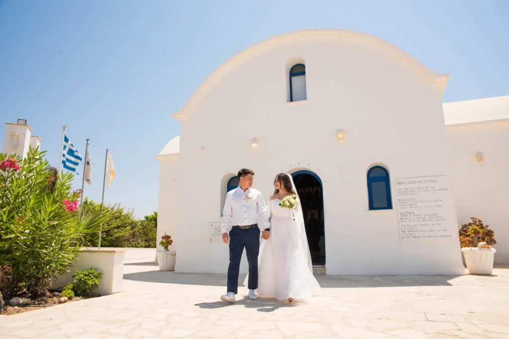 חתן וכלה הולכים יחד ליד קפלה לבנה בקפריסין במהלך חתונה רשמית לזוג מישראל
