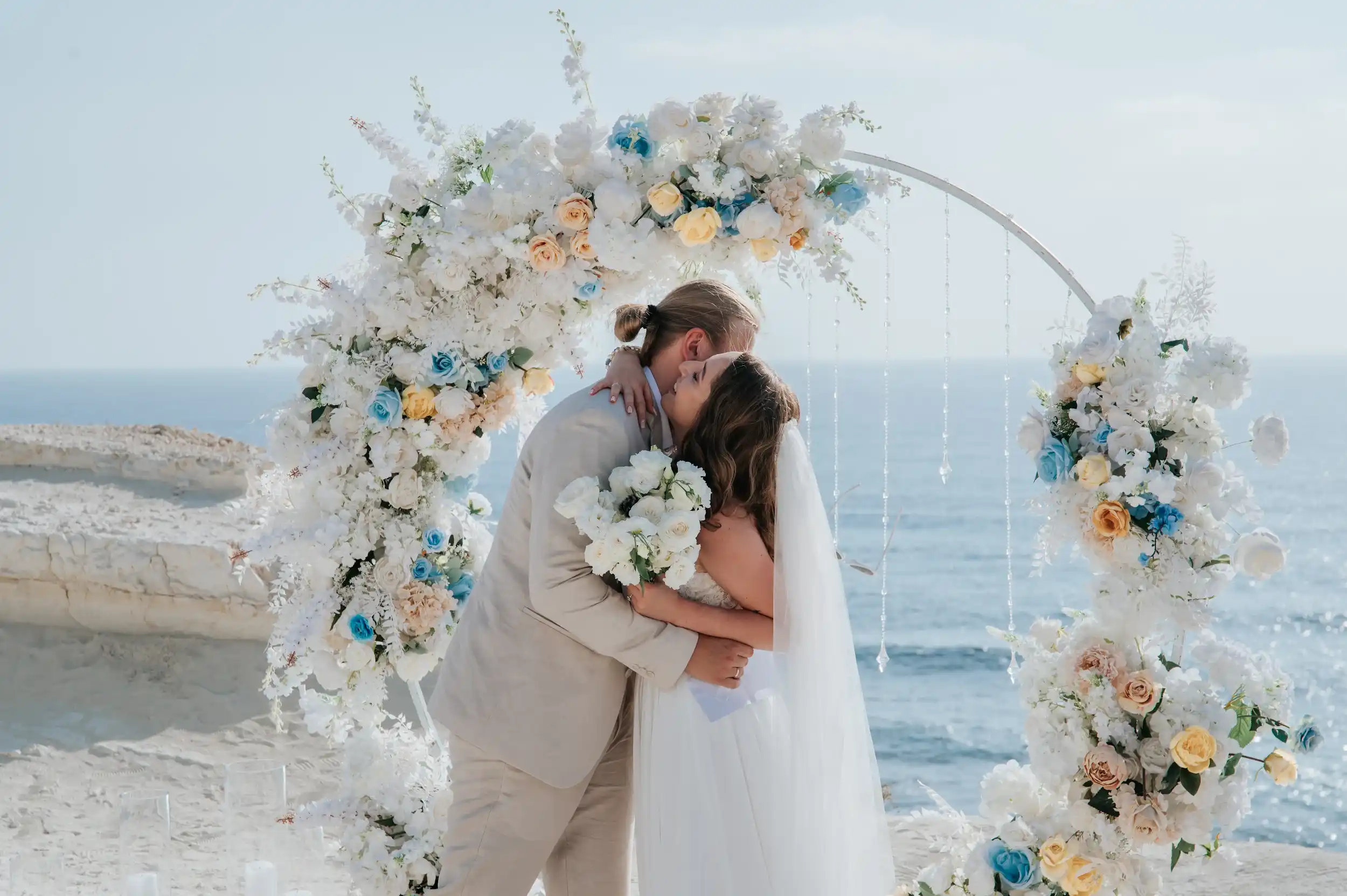 חתן וכלה מתנשקים תחת קשת פרחים ליד הים בחתונת יעד רומנטית בקפריסין לזוג מישראל