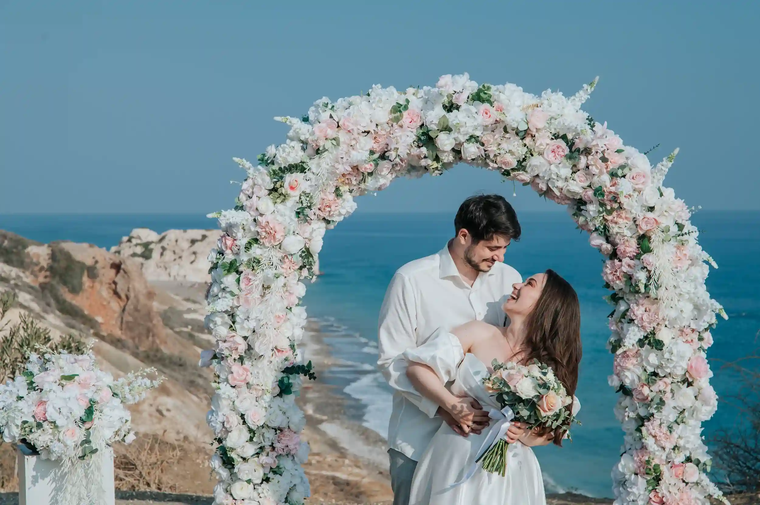 חתן וכלה מחייכים תחת קשת פרחים על רקע הים במהלך חתונת יעד רומנטית בקפריסין לזוג מישראל