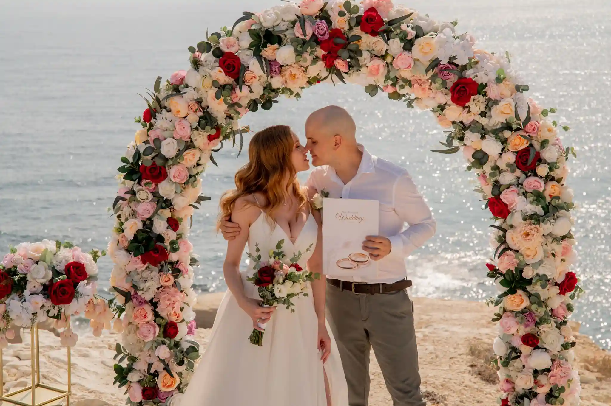 חתן וכלה מתנשקים תחת קשת פרחים עם תעודת נישואין במהלך חתונה אזרחית רשמית בקפריסין לזוג מישראל