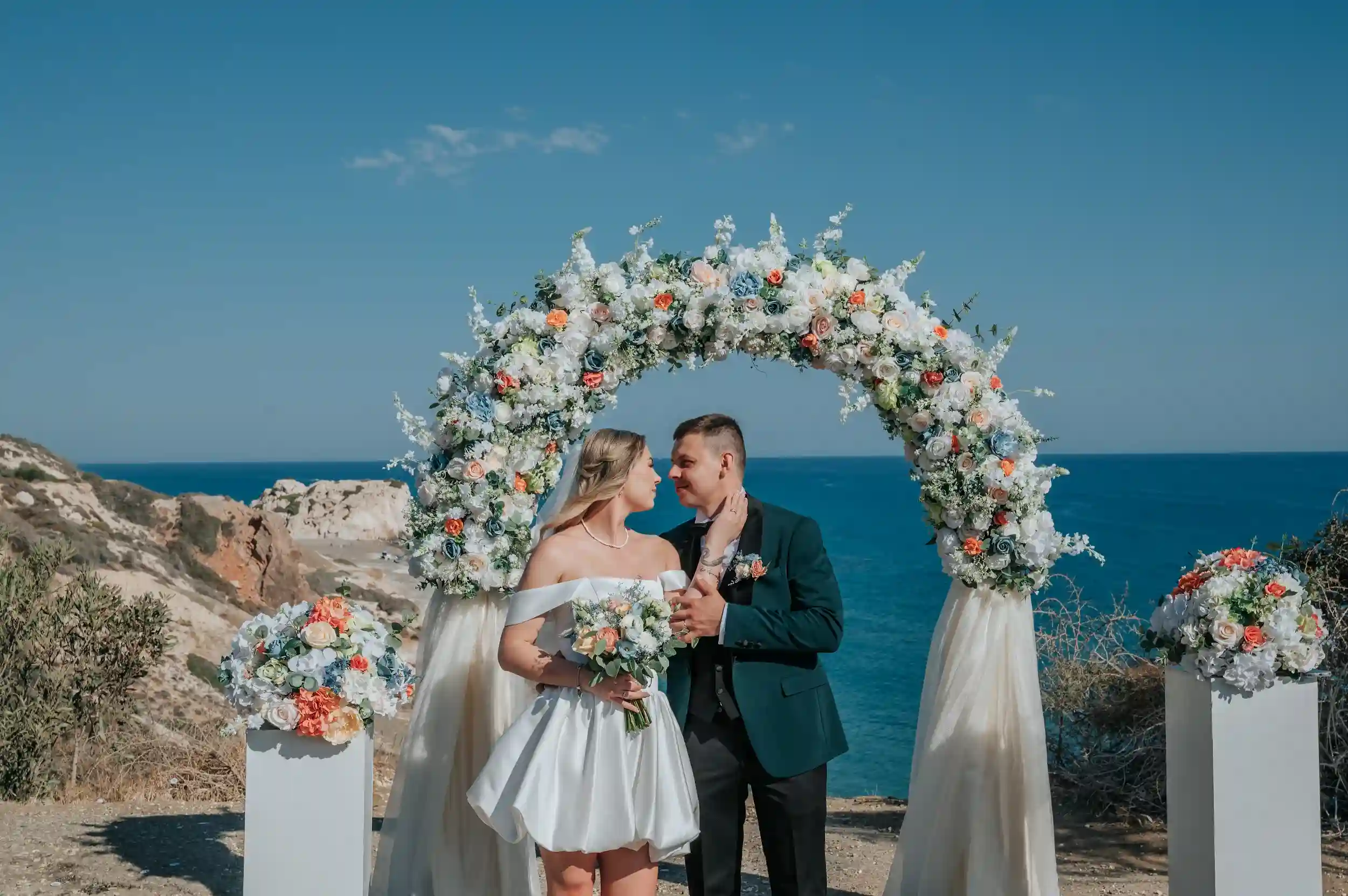 חתן וכלה עומדים תחת קשת פרחים מול הים במהלך טקס חתונה רומנטי בקפריסין לזוג מישראל