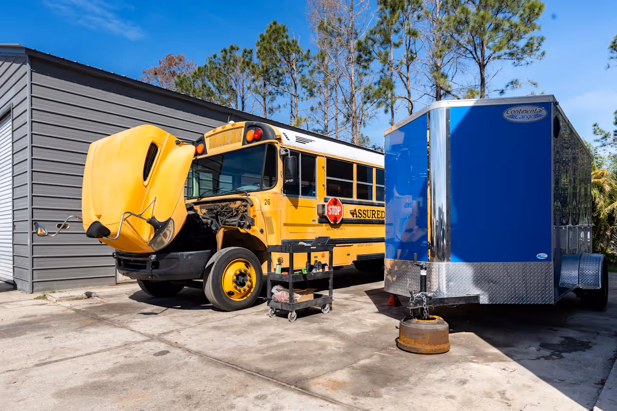 A school bus with its hood open next to a blue trailer