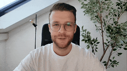 Ein lächelnder Mann mit Brille sitzt vor einem Fenster mit einem Pflanzenbaum und einer Stehlampe im Hintergrund.