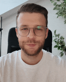 Ein Mann mit Bart und Brille sitzt vor einer Pflanze und lächelt in die Kamera.