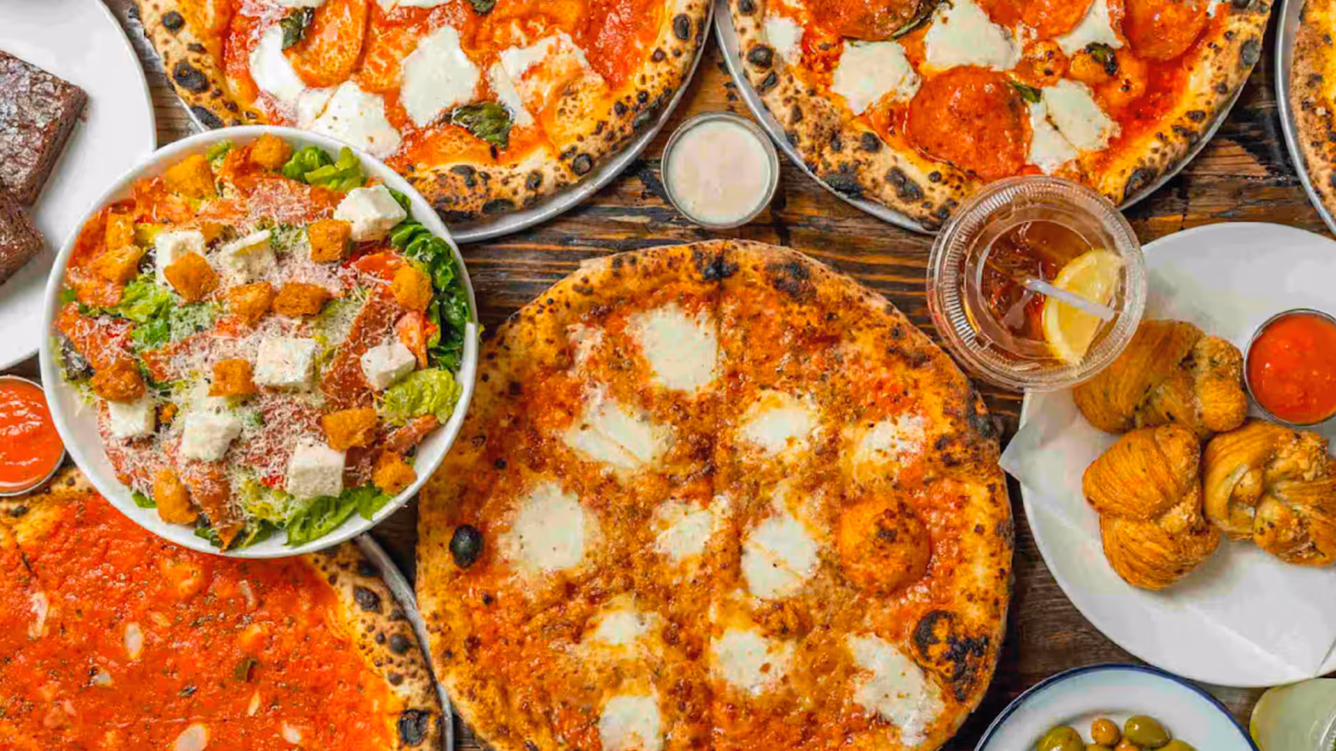 Variety of wood-fired pizzas, fresh salads, and Italian appetizers spread on a table at Roberta’s Pizza restaurant.
