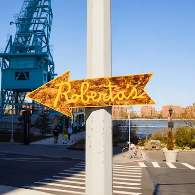 Outdoor gold arrow sign pointing towards Roberta’s Pizza restaurant with a blue crane and city skyline in the background.