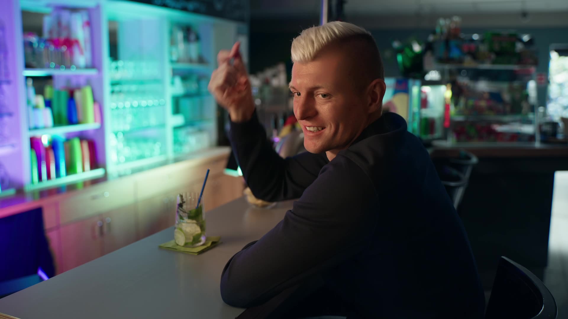 Toni Kroos sitzt lächelnd an einer Bar, blickt zur Seite und schnippt mit den Fingern.