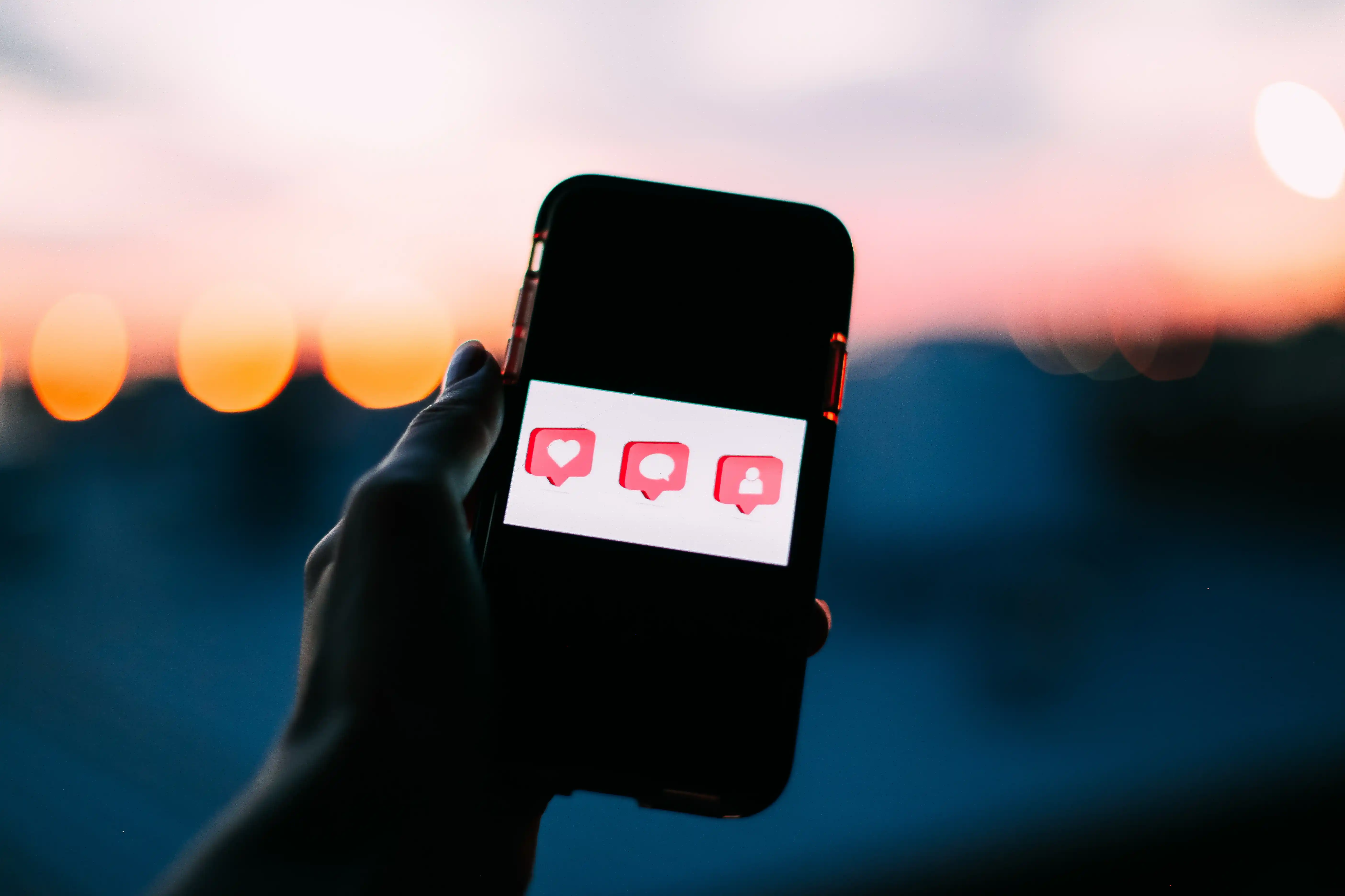 Eine Hand hält ein Smartphone mit Social-Media-Benachrichtigungssymbolen vor einem Sonnenuntergang.