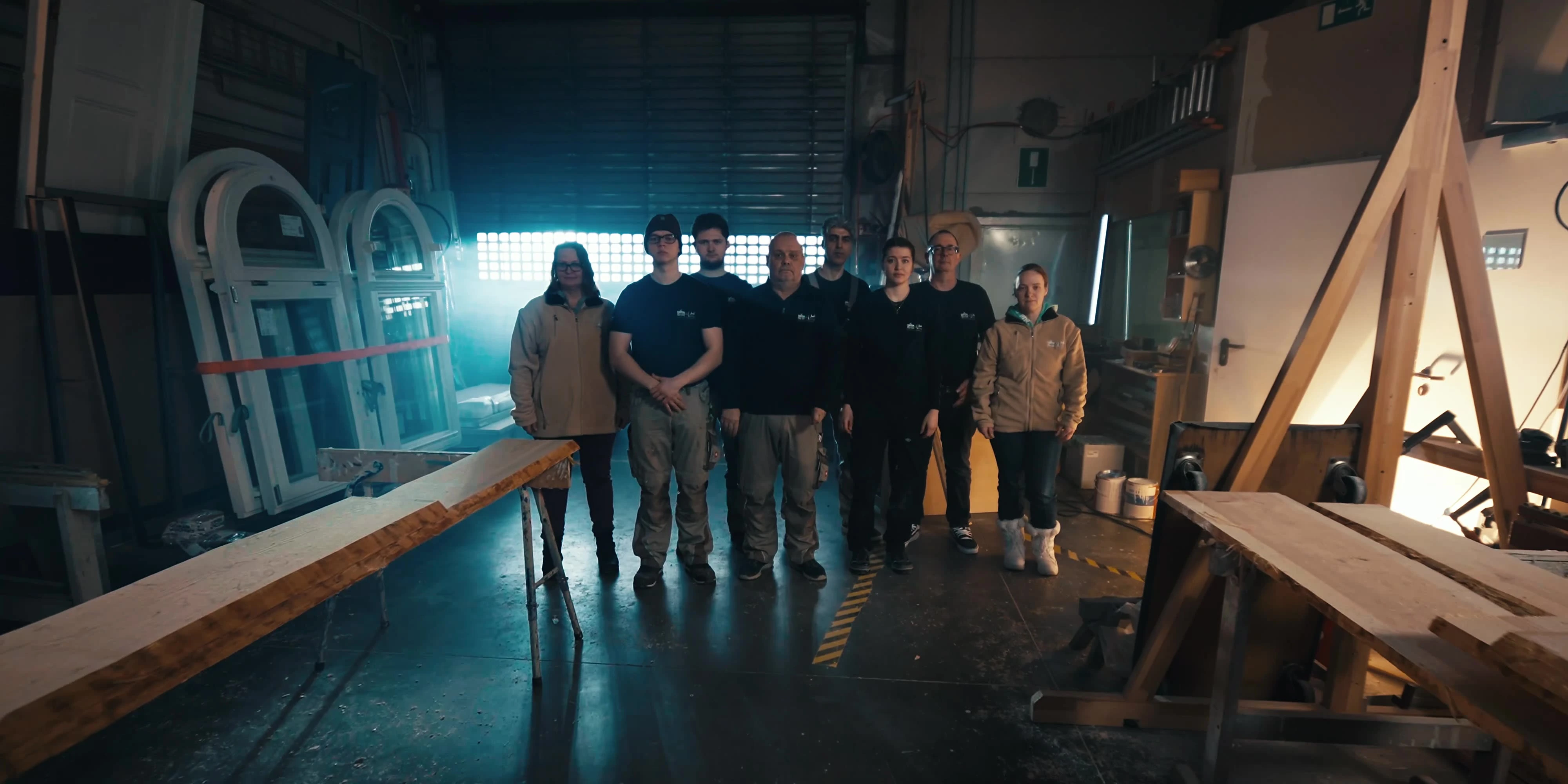 Team einer Tischlerei steht gemeinsam in der Werkstatt, zwischen Holzbohlen und fertigen Rundbogenfenstern