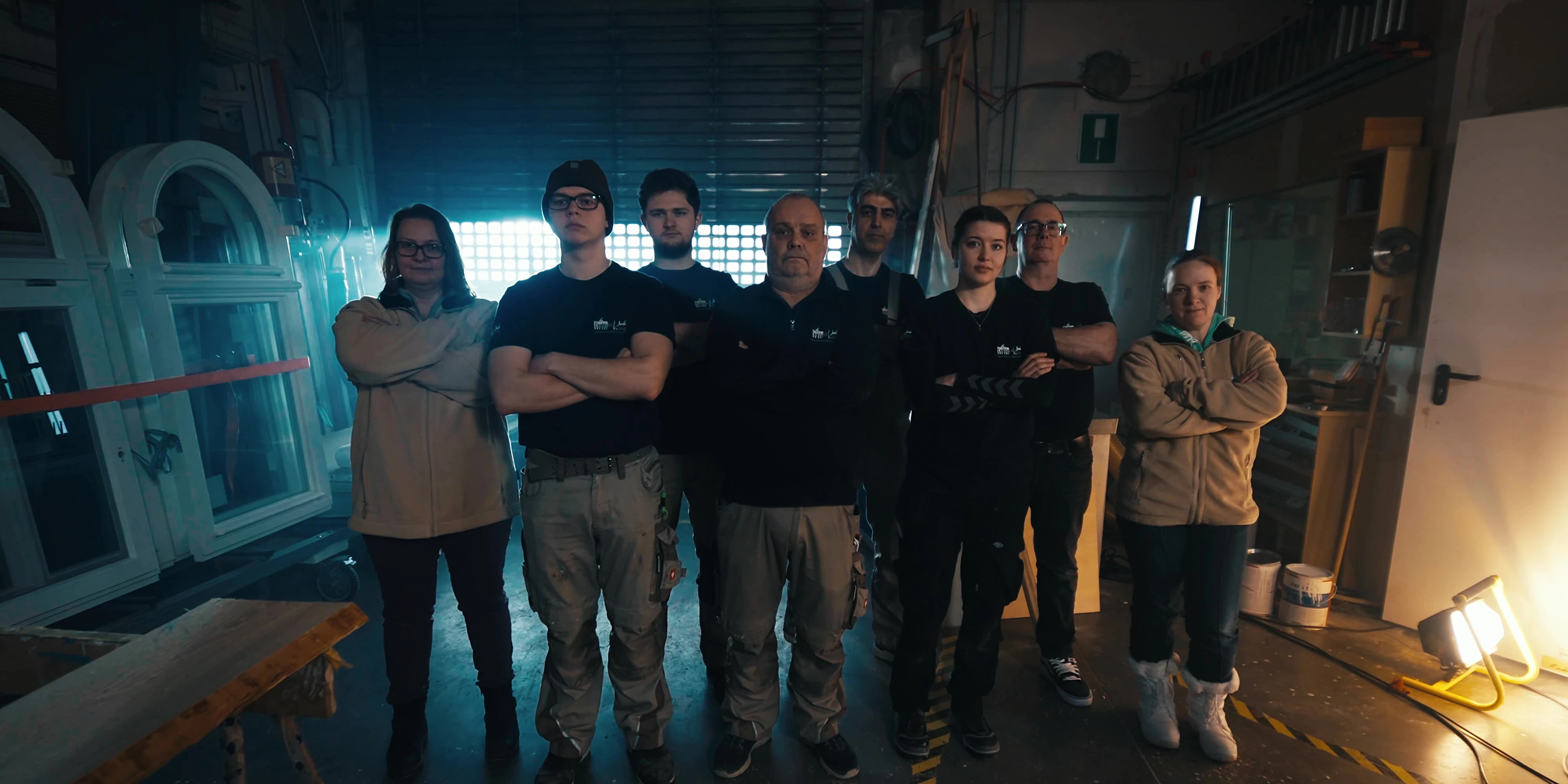 Team einer Tischlerei steht gemeinsam in der Werkstatt, zwischen Holzbohlen und fertigen Rundbogenfenstern