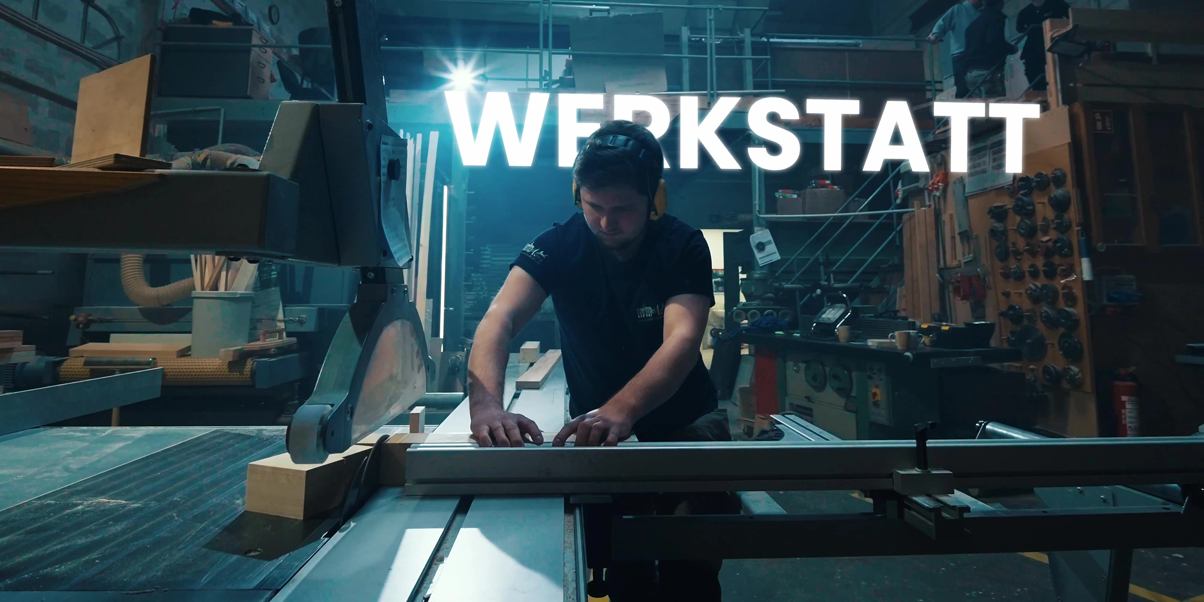 Tischler mit Gehörschutz richtet ein Werkstück an der Formatkreissäge aus, darüber der Schriftzug „Werkstatt“