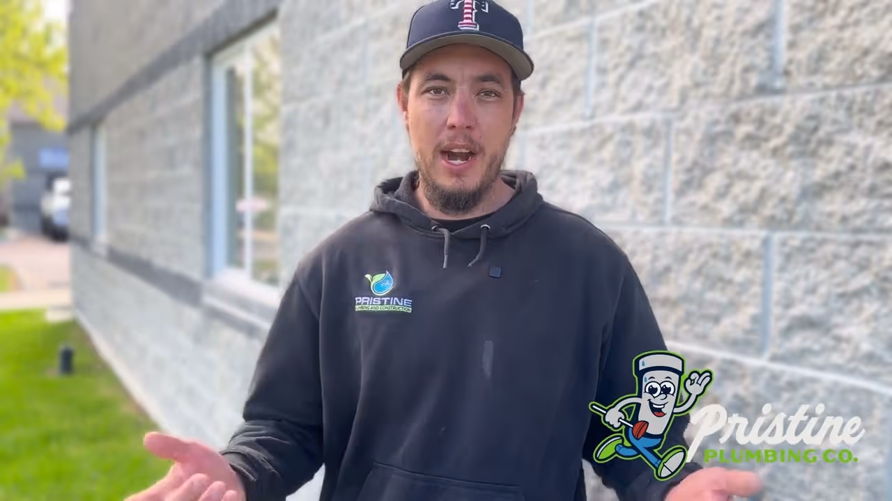 Man wearing a black Pristine Plumbing hoodie and baseball cap standing outdoors near a gray brick wall.