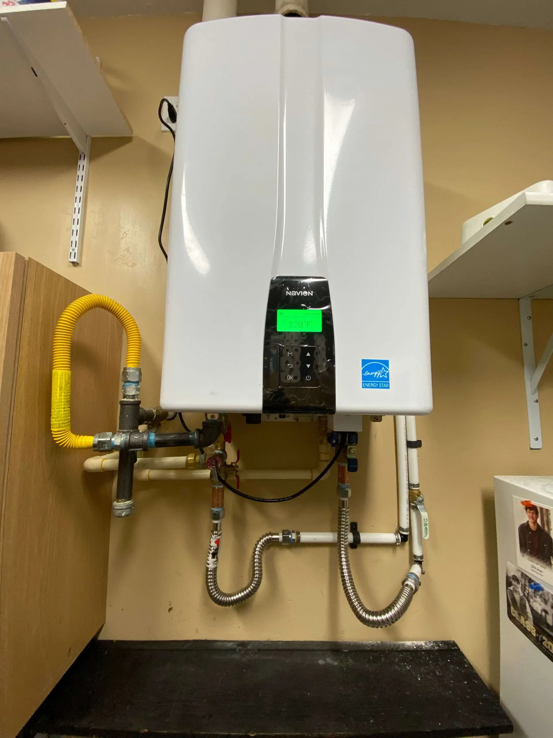 Wall-mounted white Navien tankless water heater with pipes and a digital display showing 120°F, installed in a beige utility room.