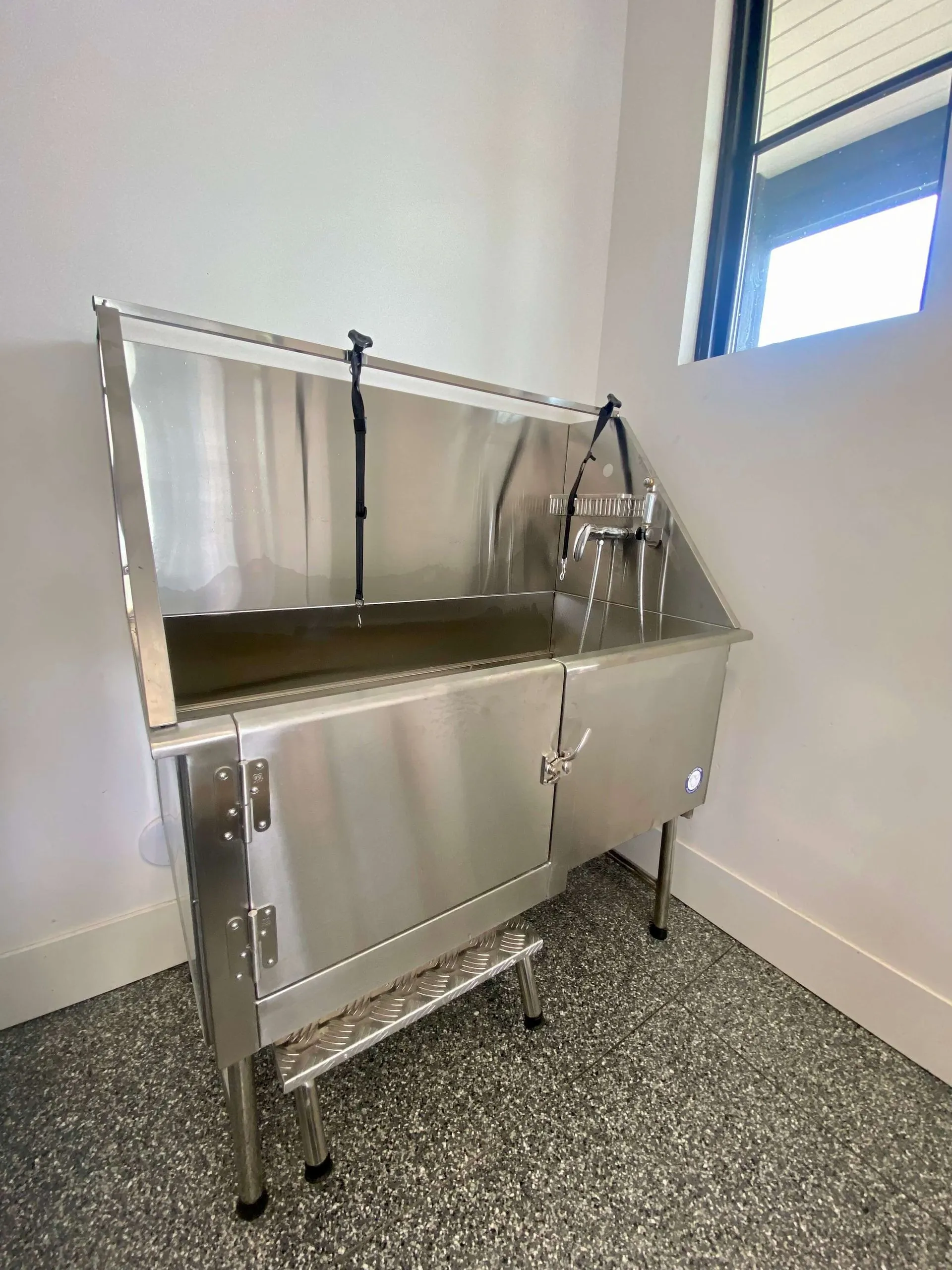 Stainless steel utility sink with a back splash, faucet, and attached hose inside a tiled room near a window.