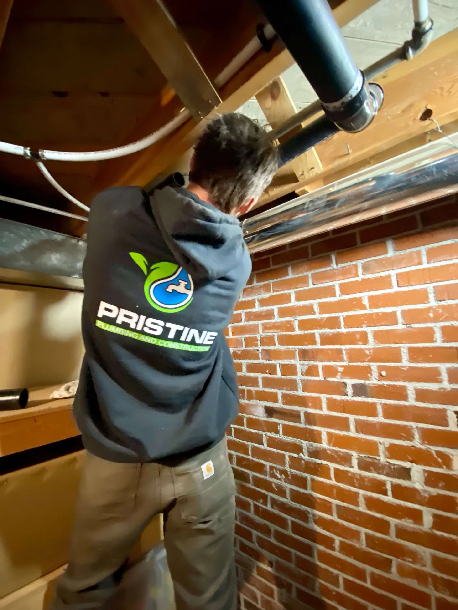 Worker in a basement wearing a Pristine Plumbing and Construction hoodie, installing or inspecting black PVC pipes near a brick wall.