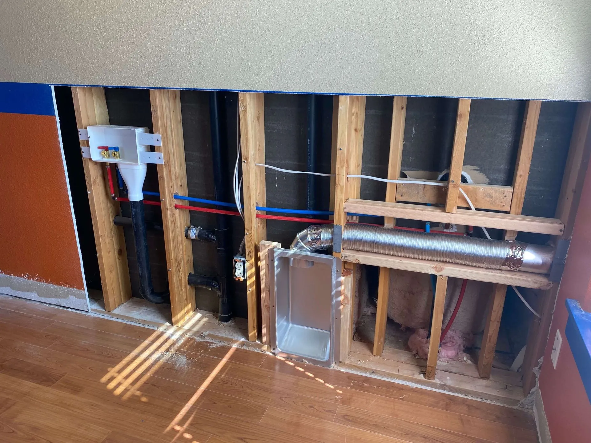 Interior wall with exposed wooden studs showing plumbing pipes, water supply lines, an electrical outlet, and a vent duct installation.