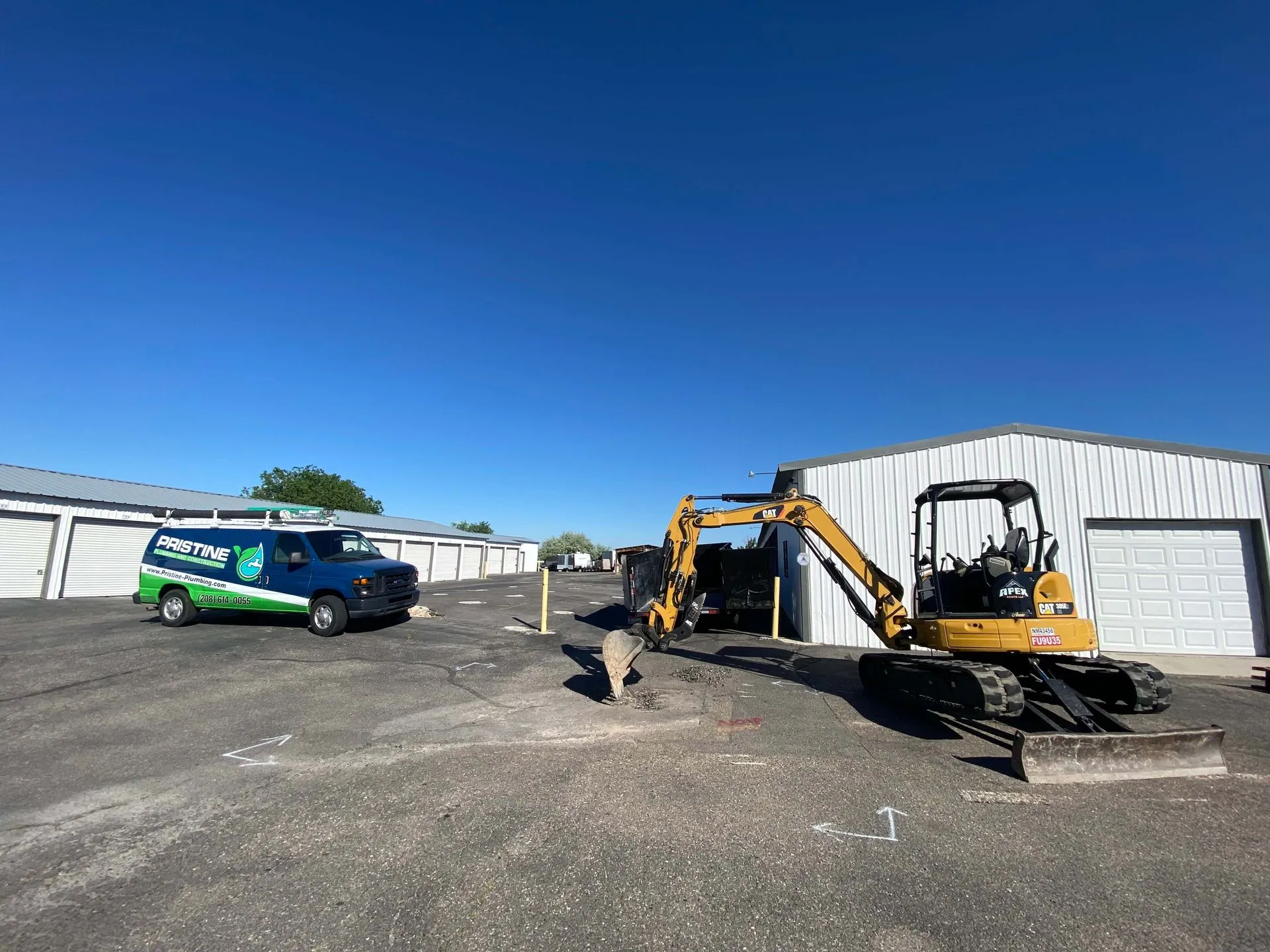 Yellow CAT mini excavator parked next to a white metal building with a blue and green Pristine Plumbing van nearby on a paved lot under a clear blue sky.