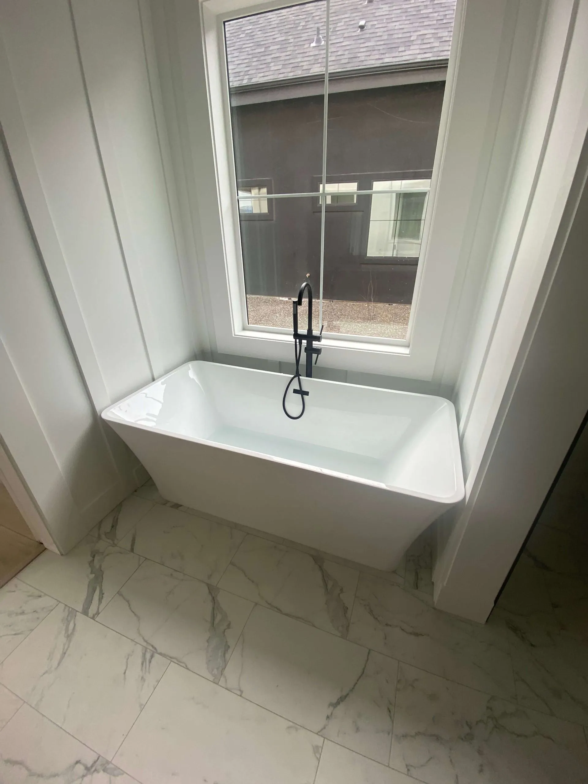 Modern white freestanding bathtub with black floor-mounted faucet under a large window with a view of a neighboring roof and wall.