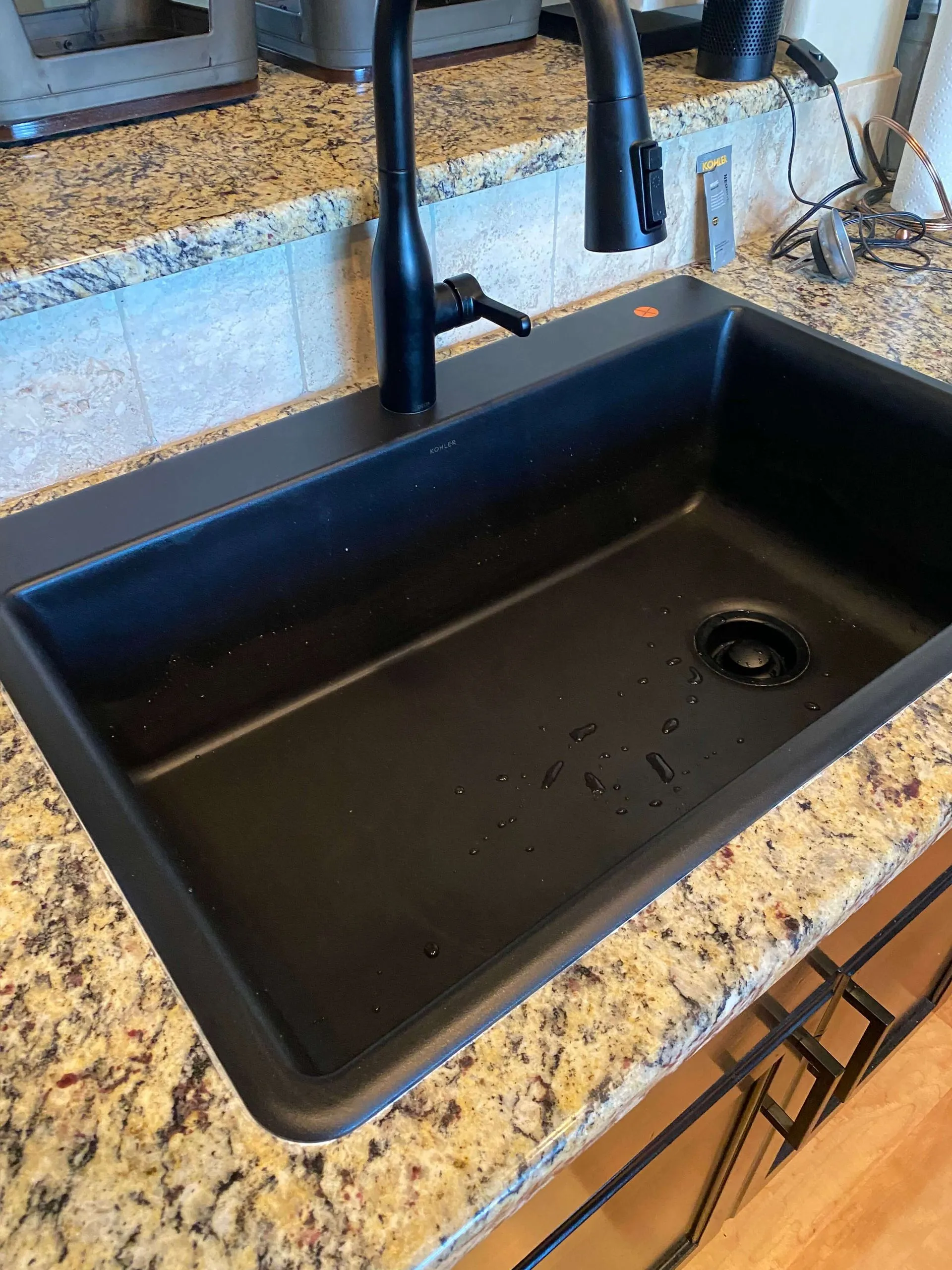 Black rectangular kitchen sink with water droplets, mounted in a granite countertop with black faucet fixtures.