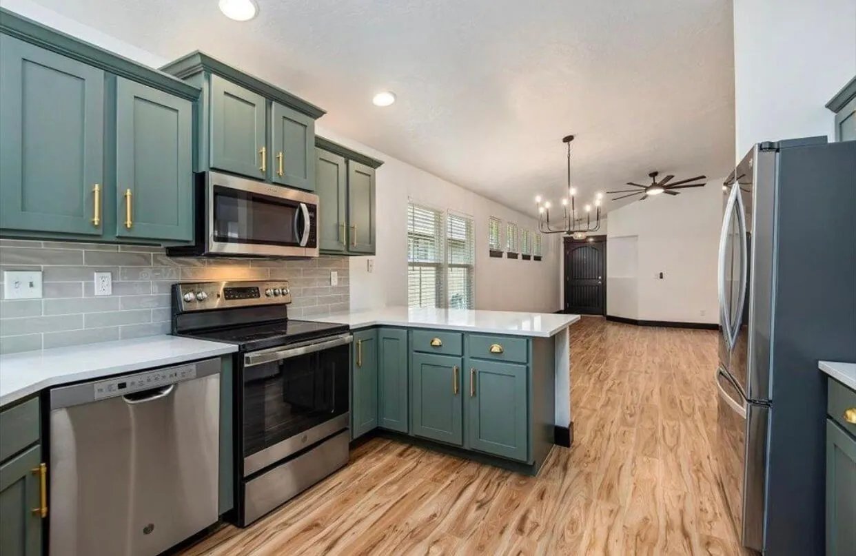 Modern kitchen with teal cabinets, stainless steel appliances, white countertops, and wood-look flooring extending to a dining area with black door and ceiling fan.