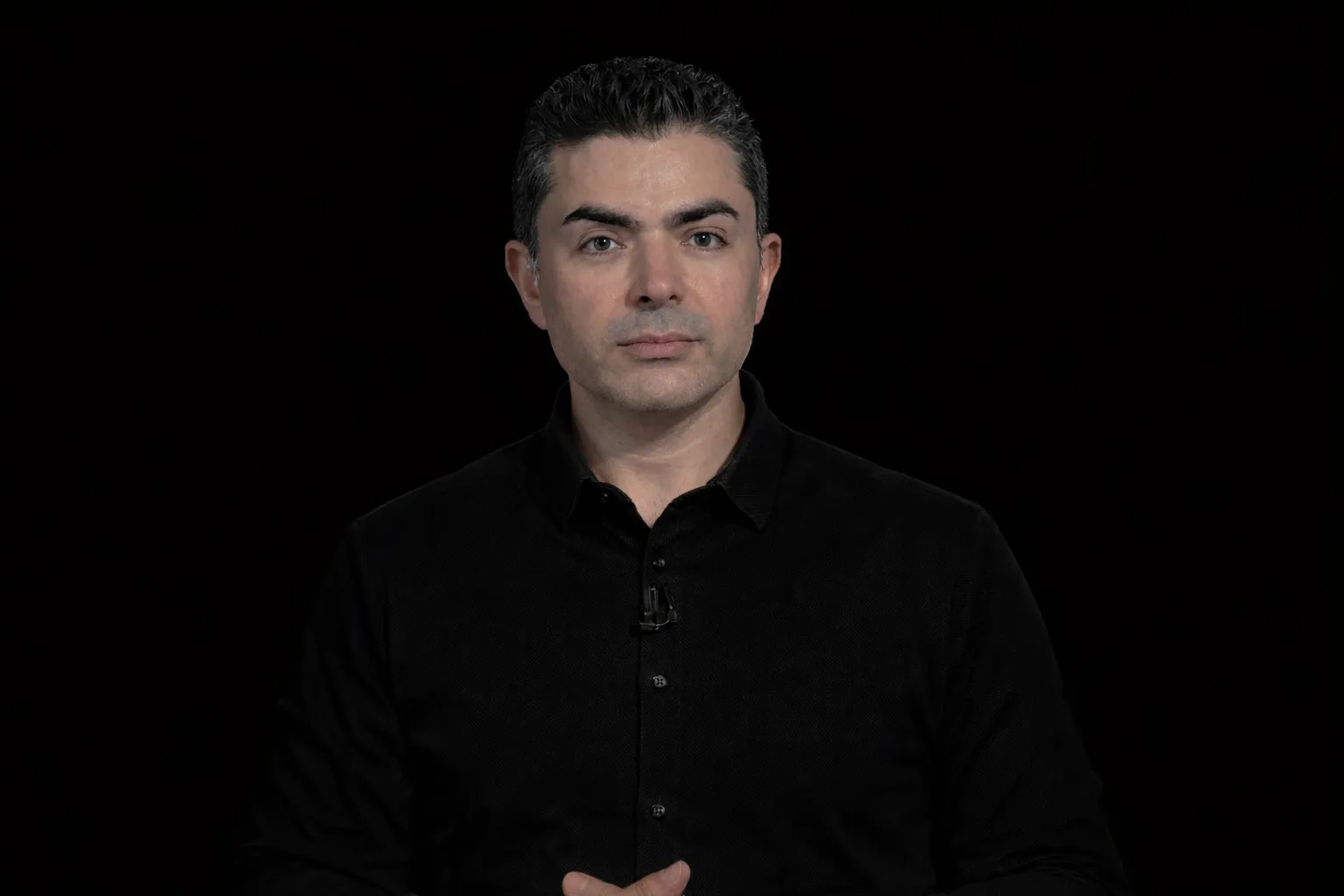 Man with dark hair wearing a black button-up shirt against a black background.