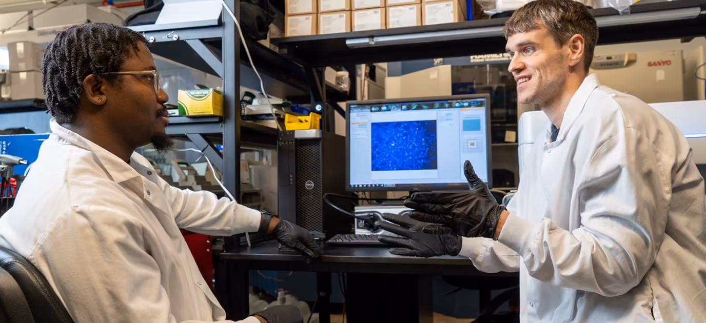 Two scientists in white lab coats and black gloves discuss data shown on a computer screen displaying a blue microscopic image in a laboratory filled with equipment and shelves.