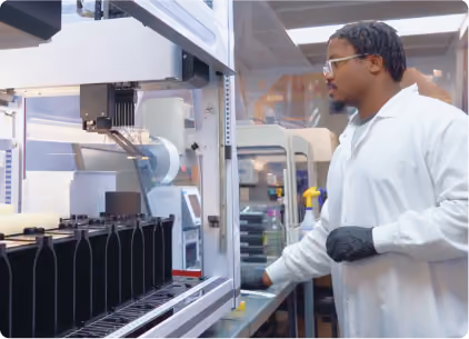 A man wearing a white lab coat and black gloves operates a large automated machine in a laboratory or manufacturing setting.
