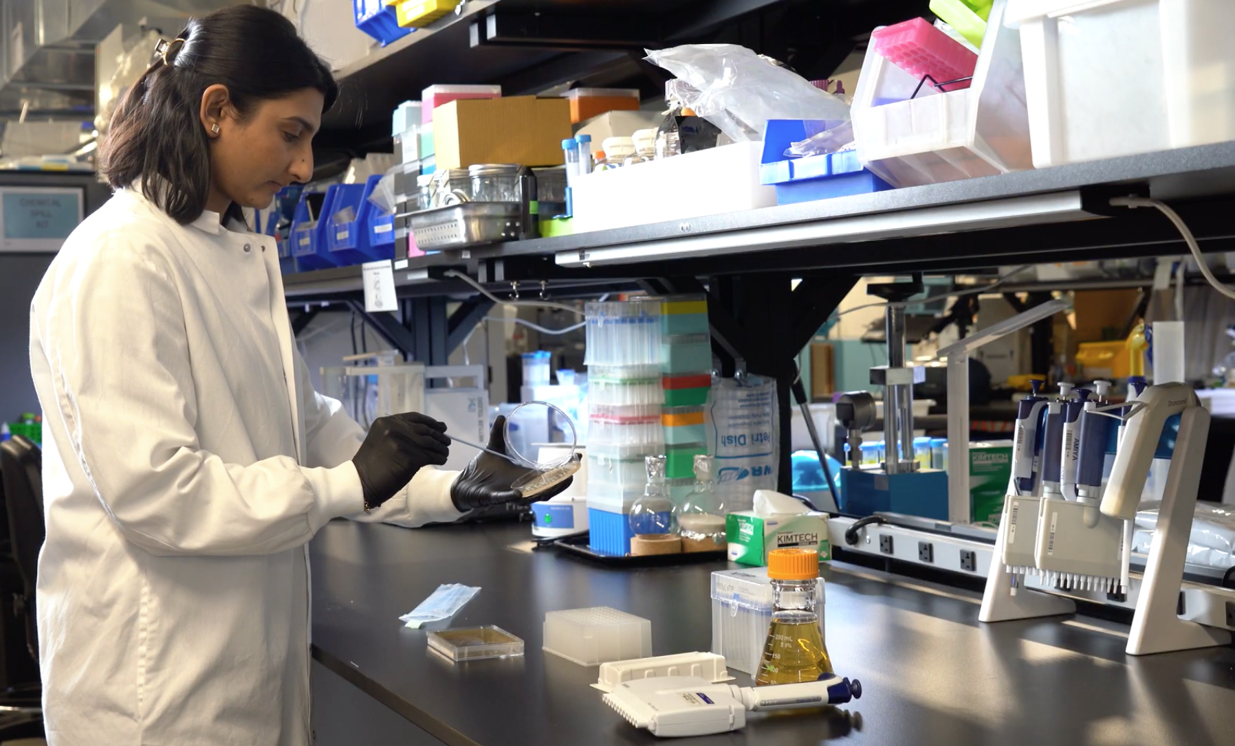 Scientist wearing a white lab coat and black gloves working in a laboratory, holding a petri dish and using a tool, surrounded by lab equipment and supplies on a cluttered black workbench.