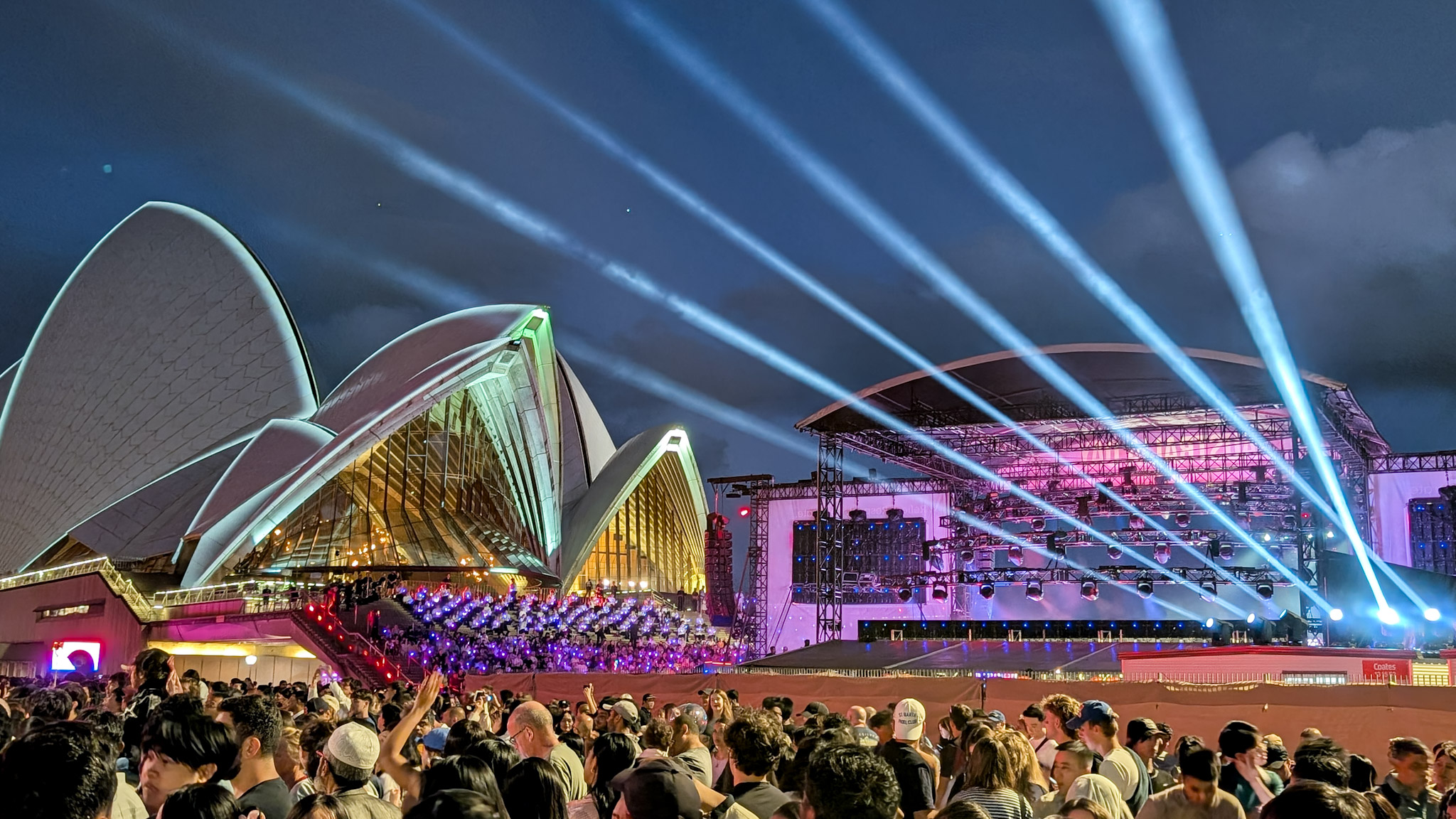 Crowd gathered at night outside Sydney Opera House with colorful stage lights and beams in the sky.