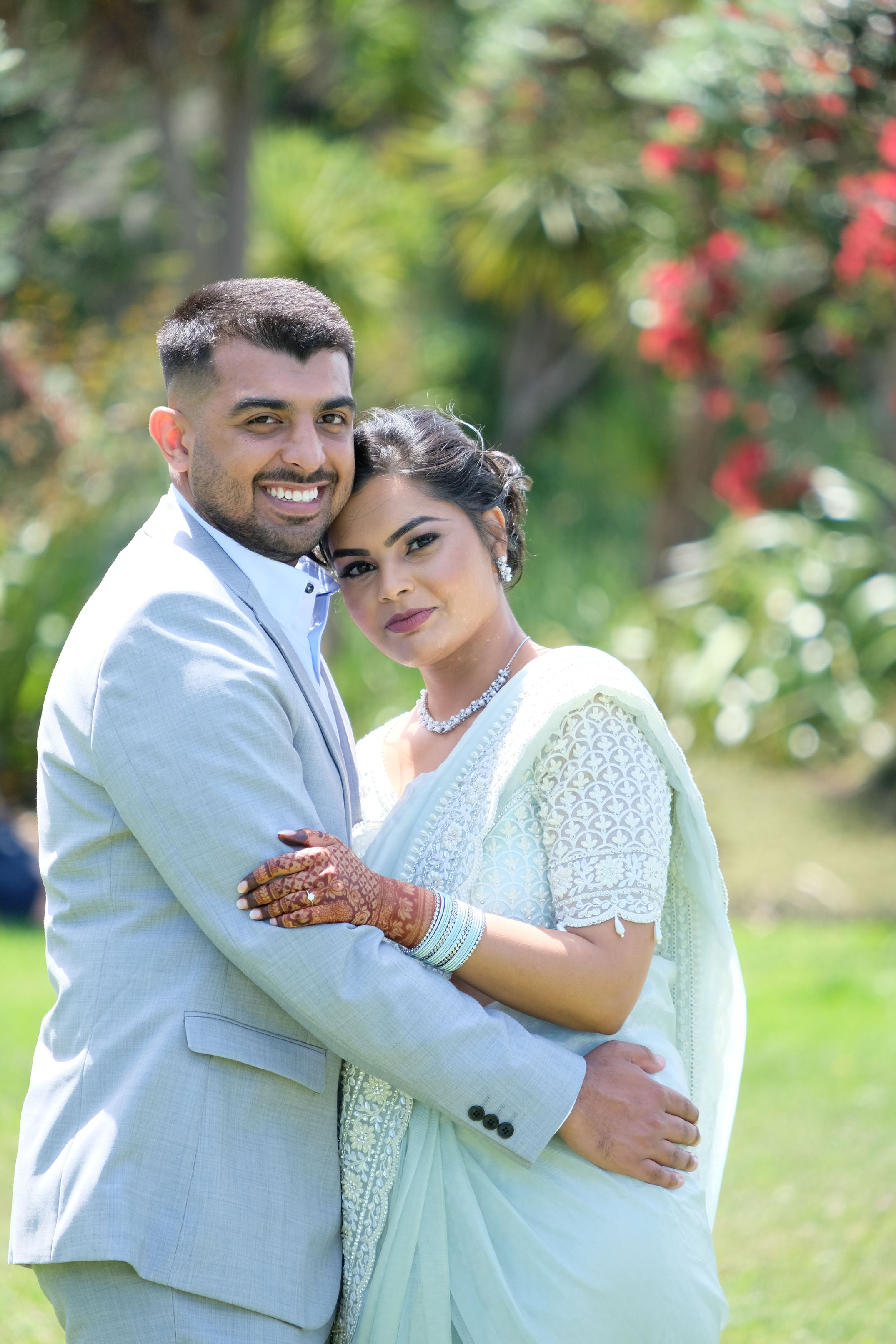 Smiling couple in formal attire embracing outdoors with greenery and flowers in background.