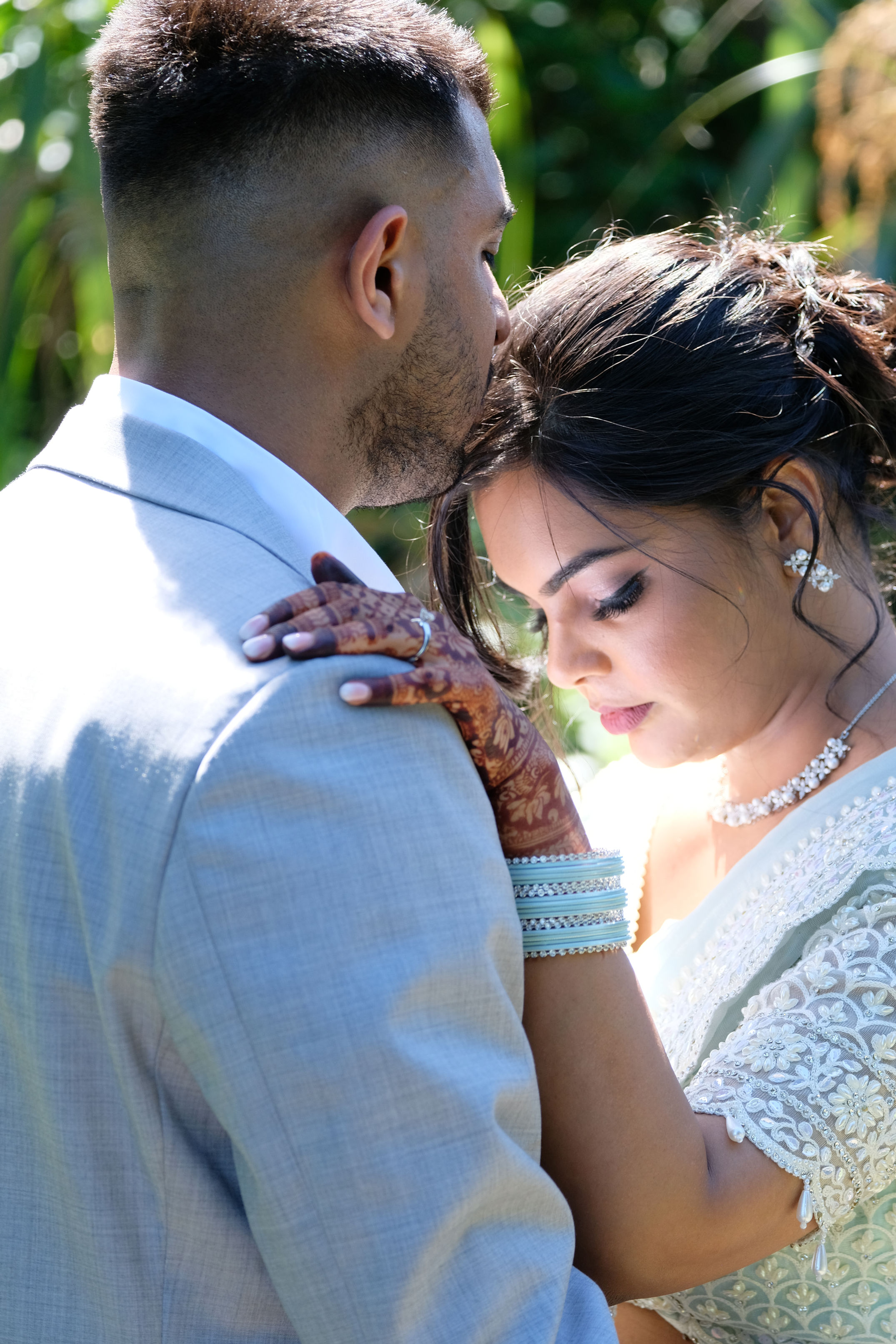 Bride in traditional henna and jewelry resting her head on groom's shoulder, who is wearing a light gray suit, outdoors.