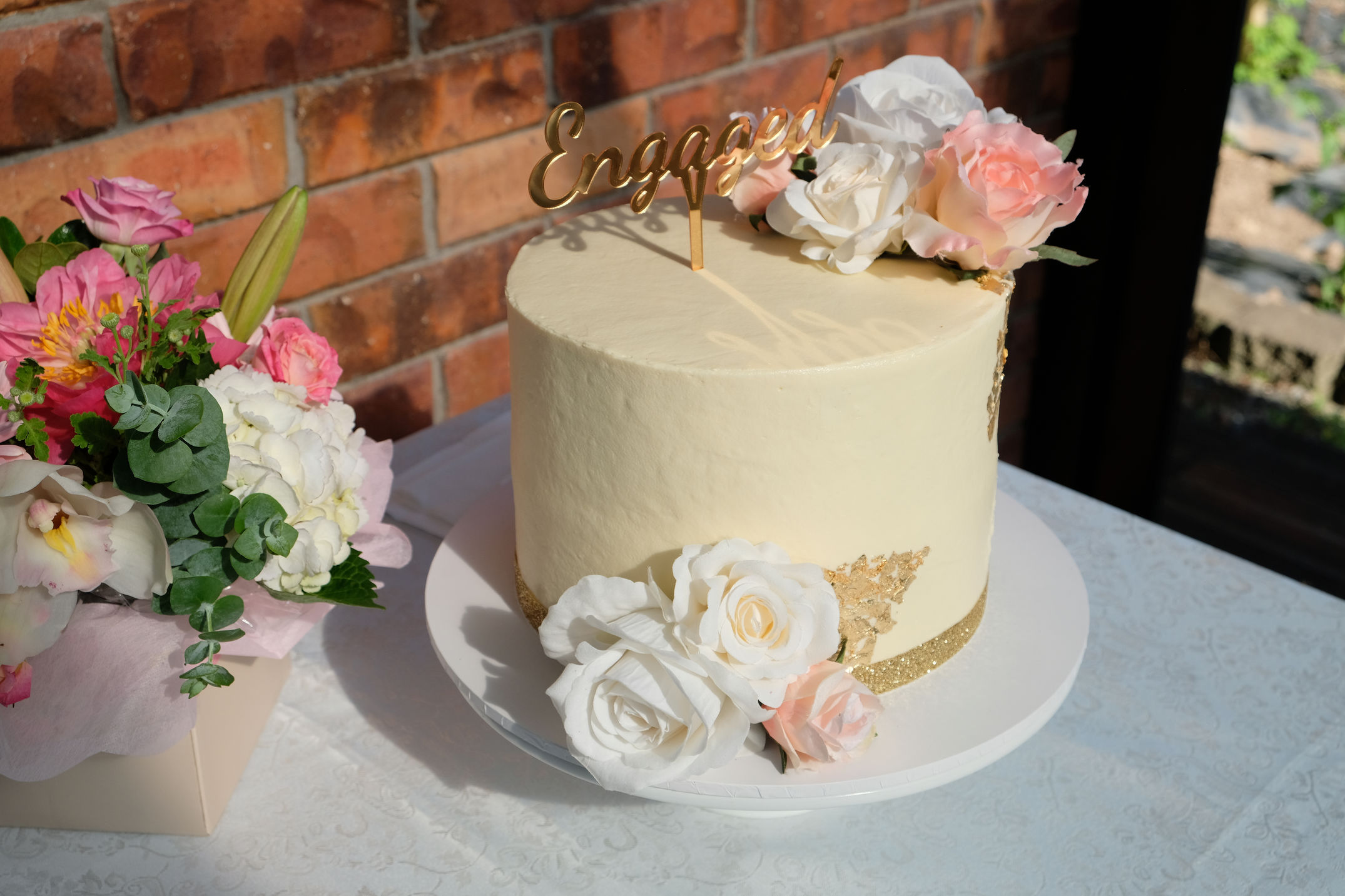 Round engagement cake with smooth cream frosting, decorated with white and pink roses and a gold glitter band, topped with a gold 'Engaged' sign.