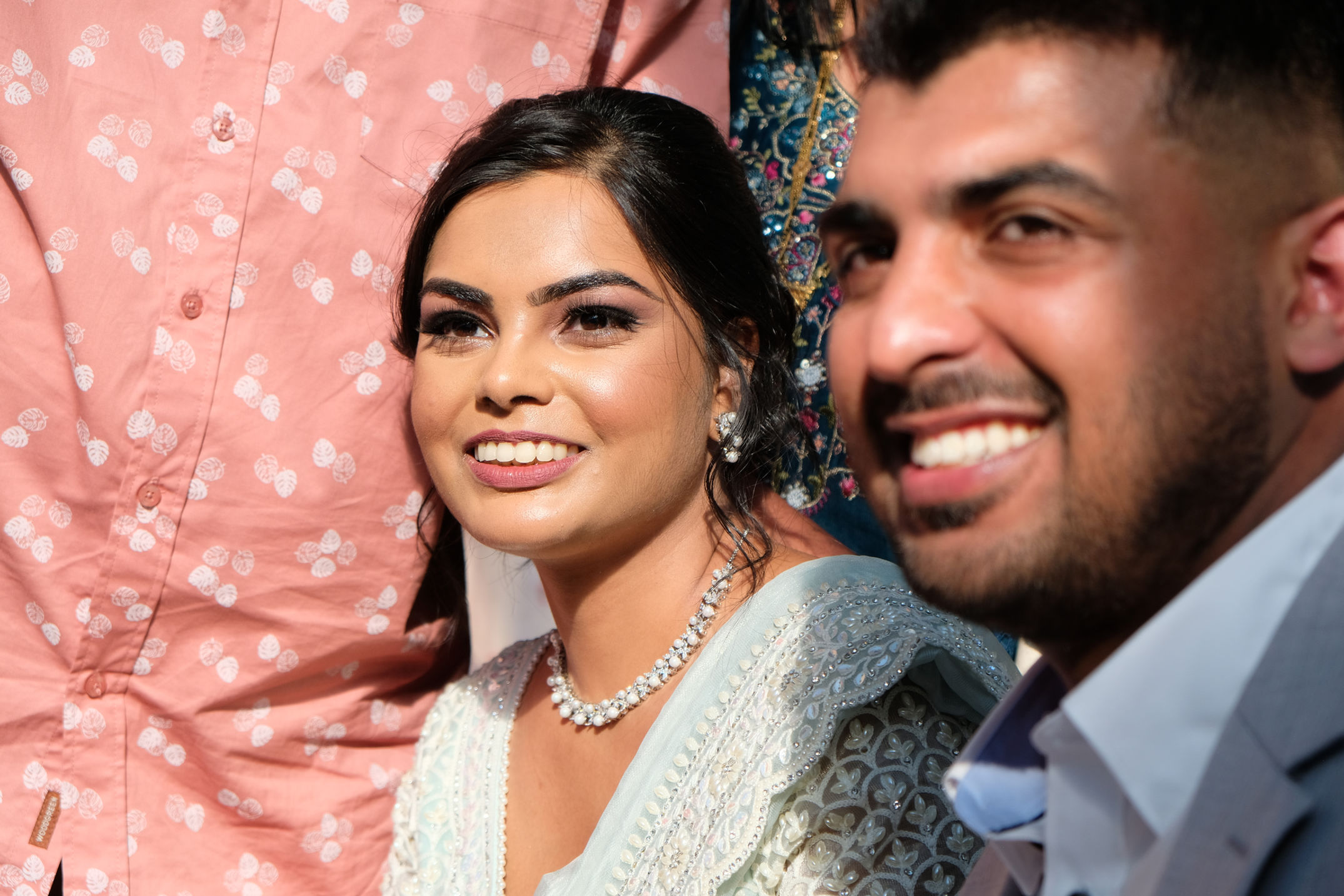 Close-up of a smiling woman dressed in a traditional light blue embellished outfit with pearl jewelry, beside a man in a gray suit smiling.