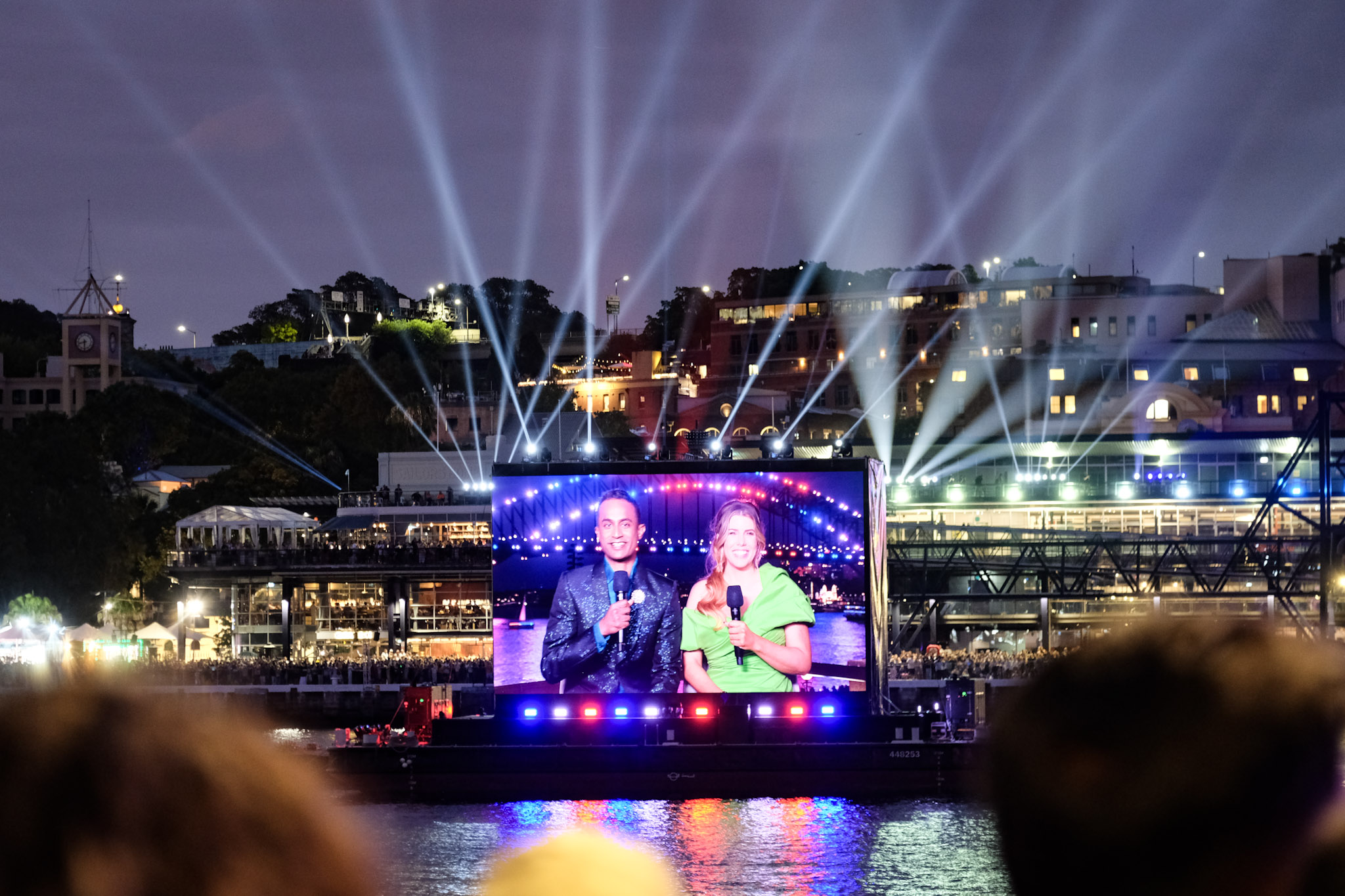 Large outdoor screen over water showing two hosts with microphones during a nighttime event with light beams above.