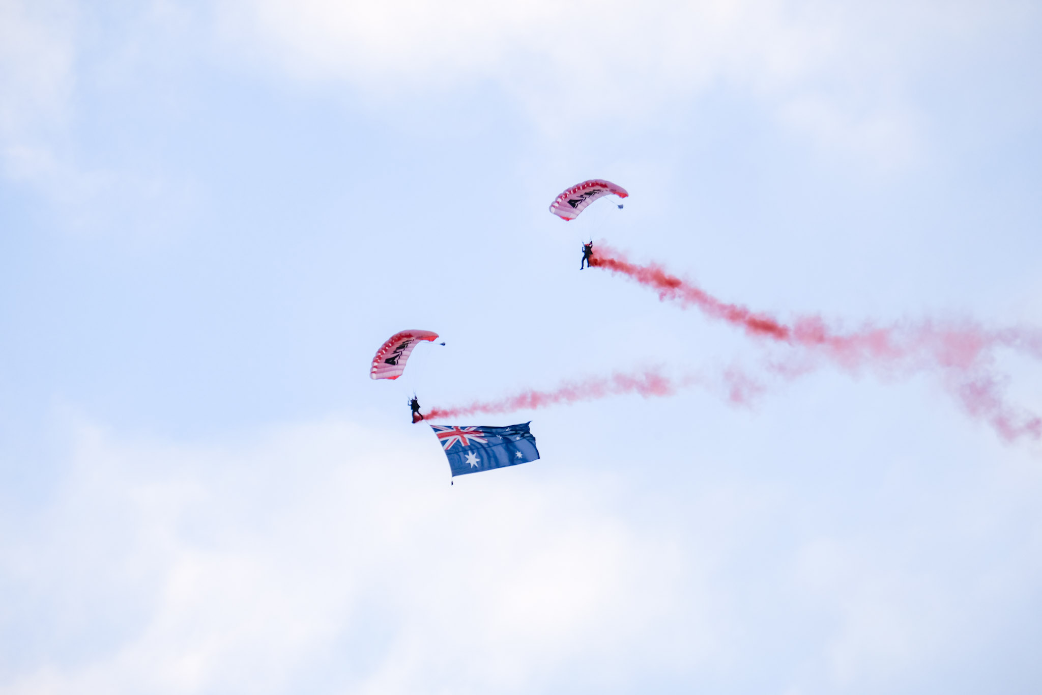 Two paragliders in the sky trailing red smoke, one carrying a large Australian flag.