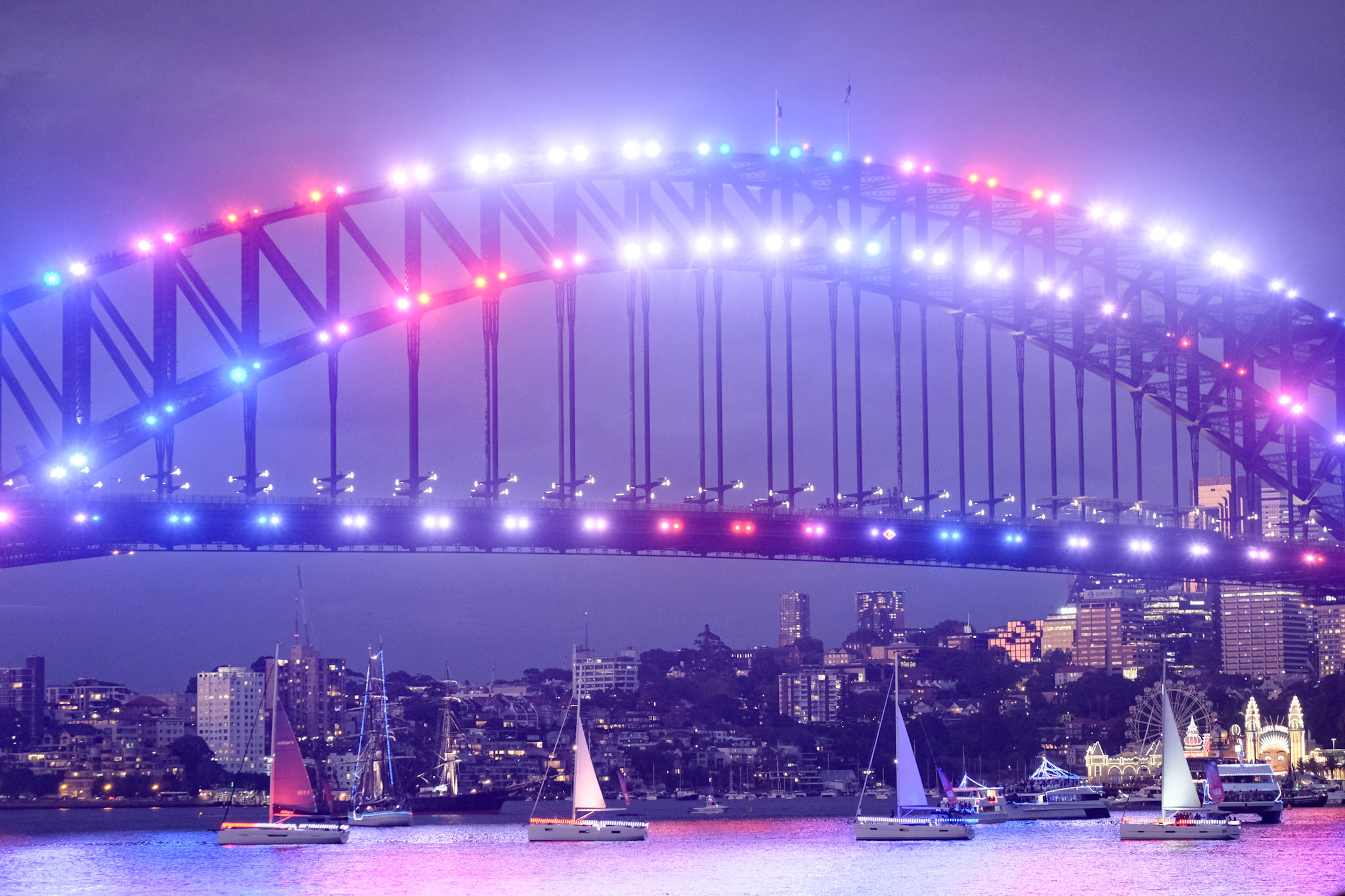 Sydney Harbour Bridge lit with colorful lights at night over water with sailboats and city skyline.
