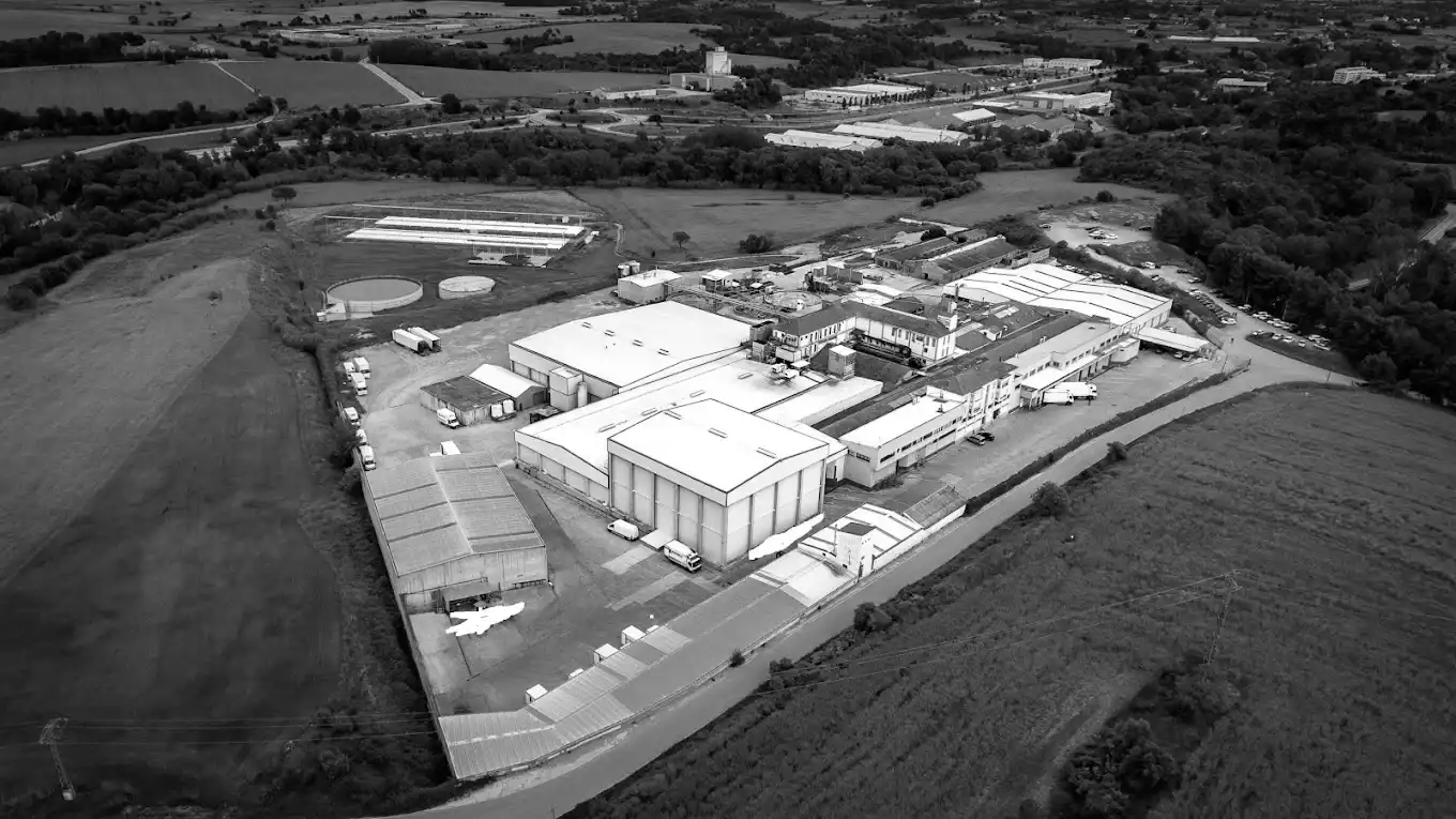 Aerial view of an industrial complex with multiple large white-roofed buildings surrounded by fields and trees.