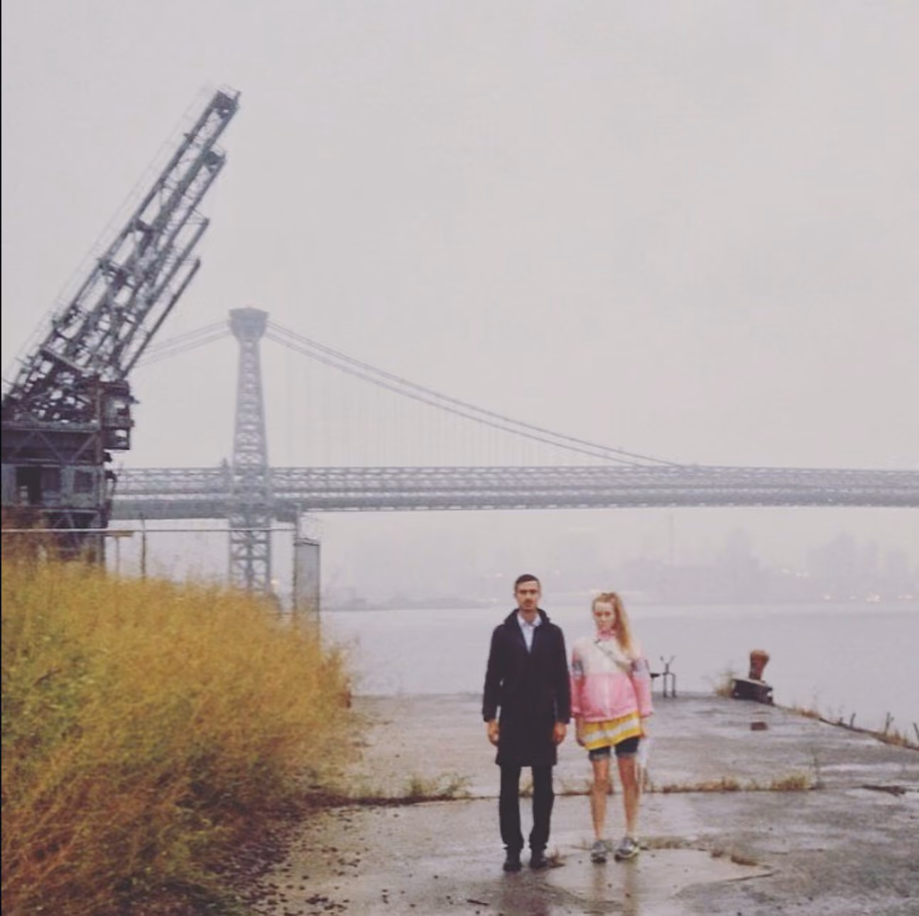 Two people standing on a concrete pier by the East River in fog, with a derelict crane and the Williamsburg Bridge behind them.