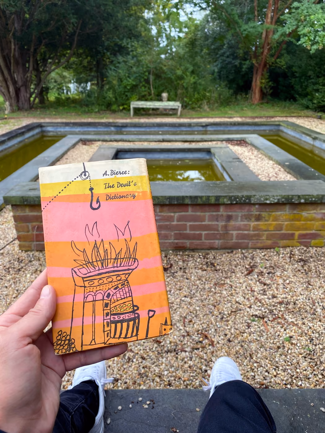 A hand holding a striped mid-century dust jacket of Ambrose Bierce's The Devil's Dictionary above a formal sunken water garden.