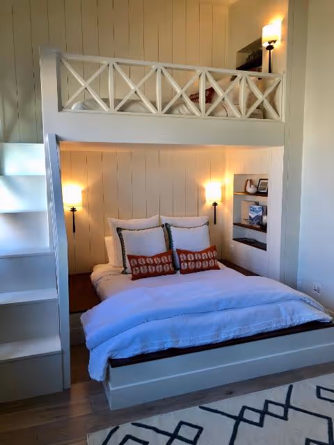 Cozy bedroom with a loft bed above, lit by wall sconces. White bedding and pillows add comfort. Built-in shelves hold books.