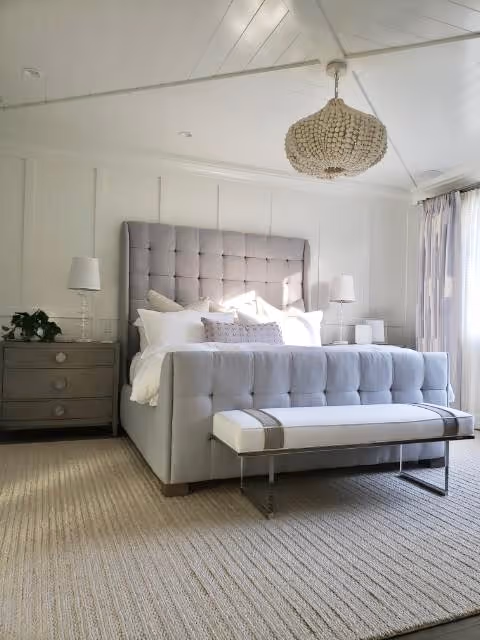A cozy bedroom with a tufted bed, soft lighting, and a woven chandelier. Sunlight streams through a window, illuminating the space.