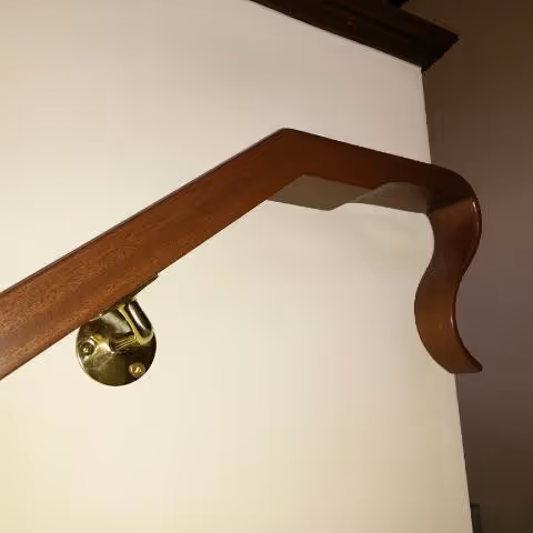 Close-up of a curved wood stair handrail with decorative volute end and brass wall bracket mount
