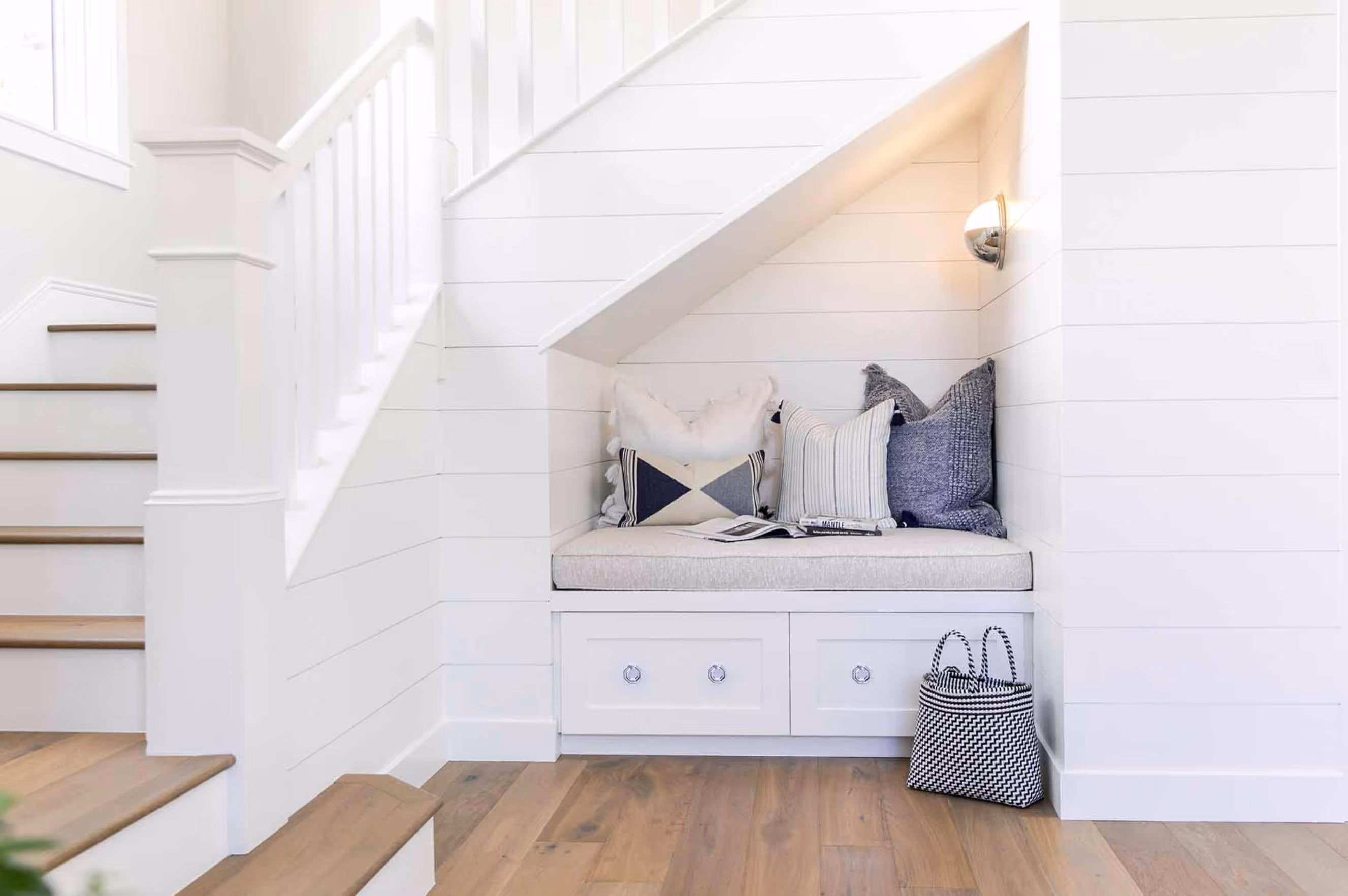 Cozy nook under stairs with cushions, soft lighting, and a basket on wooden floor, creating a serene reading space.