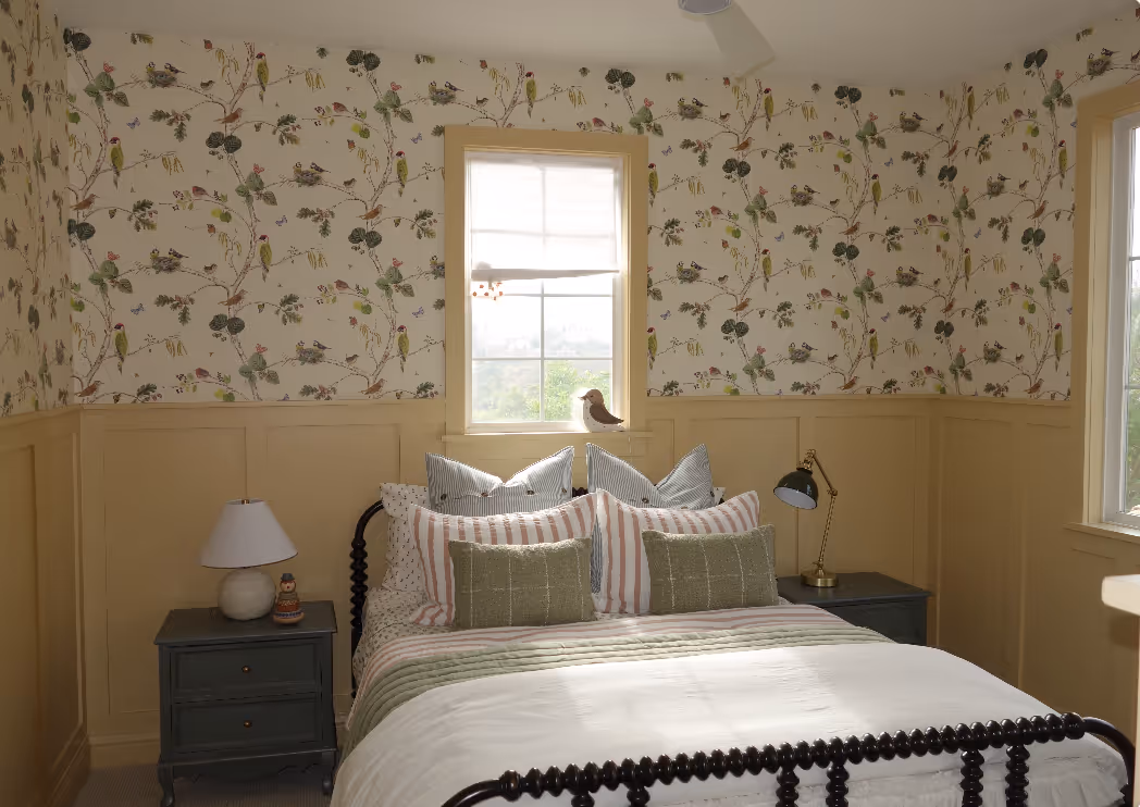 Cozy bedroom with bird-themed wallpaper, a window, and a neatly made bed with striped and green pillows, flanked by nightstands.